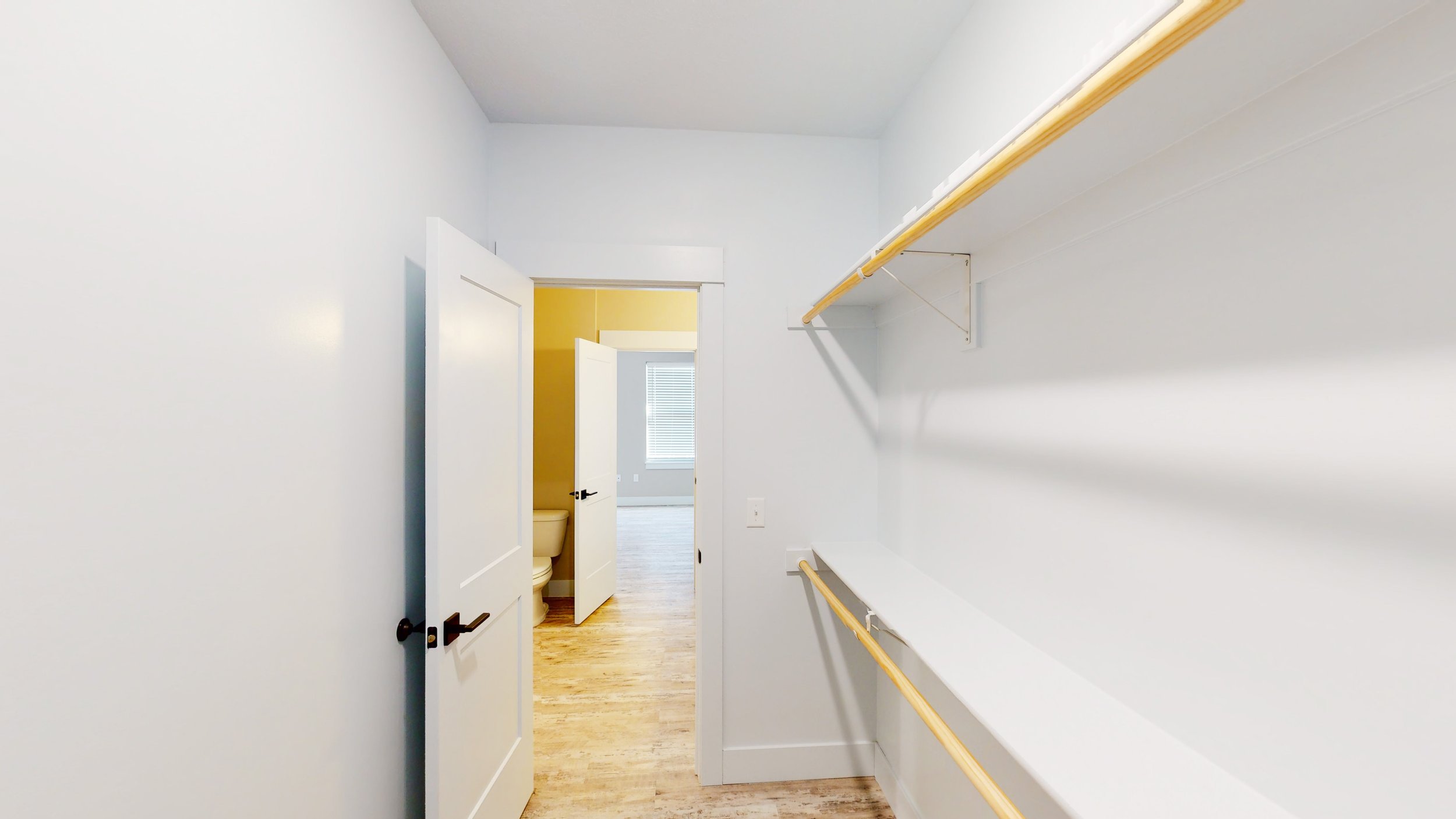 Empty walk-in closet with white walls, wooden shelves, and hangers, adjacent to a room with hardwood flooring and a visible bathroom.