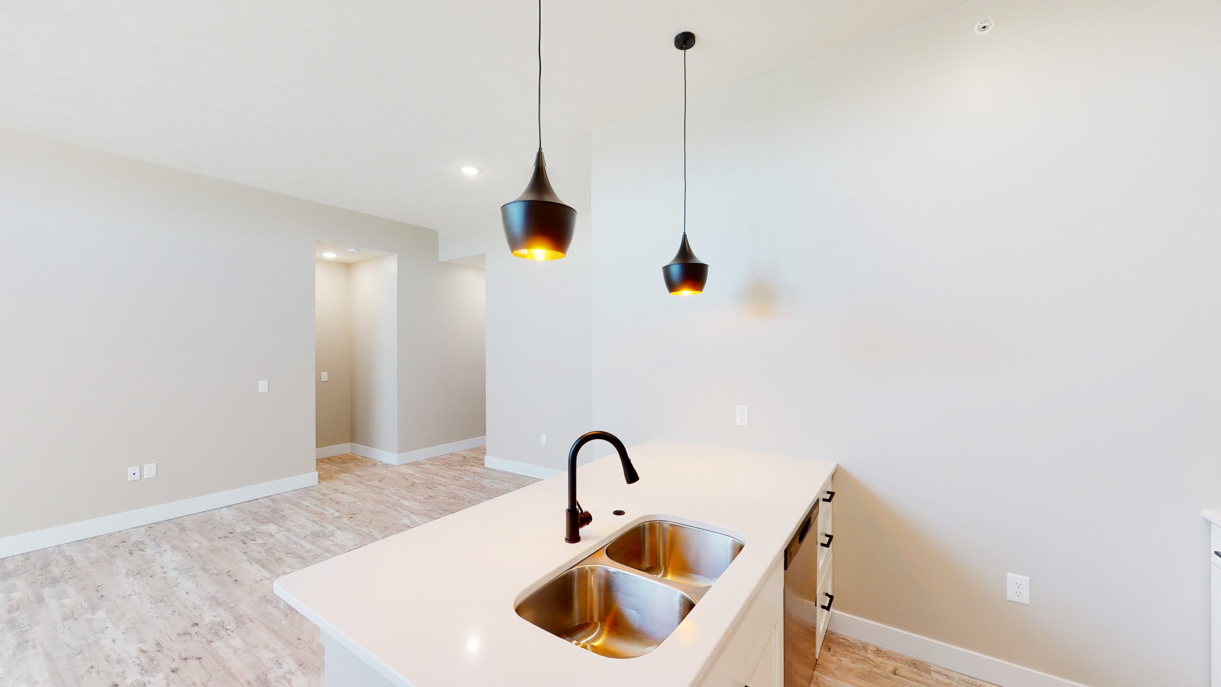Modern kitchen interior with white countertops, stainless steel sink, black faucet, wood flooring, and pendant lighting.
