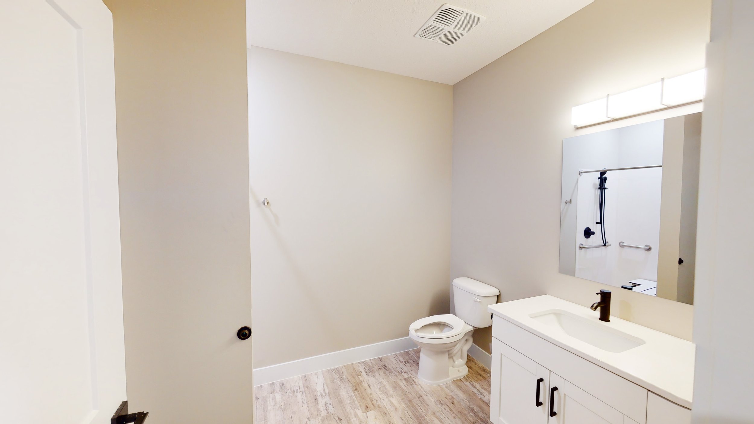 Modern bathroom with white toilet, vanity with sink, large mirror, wall light, towel bar, and wood-style flooring.