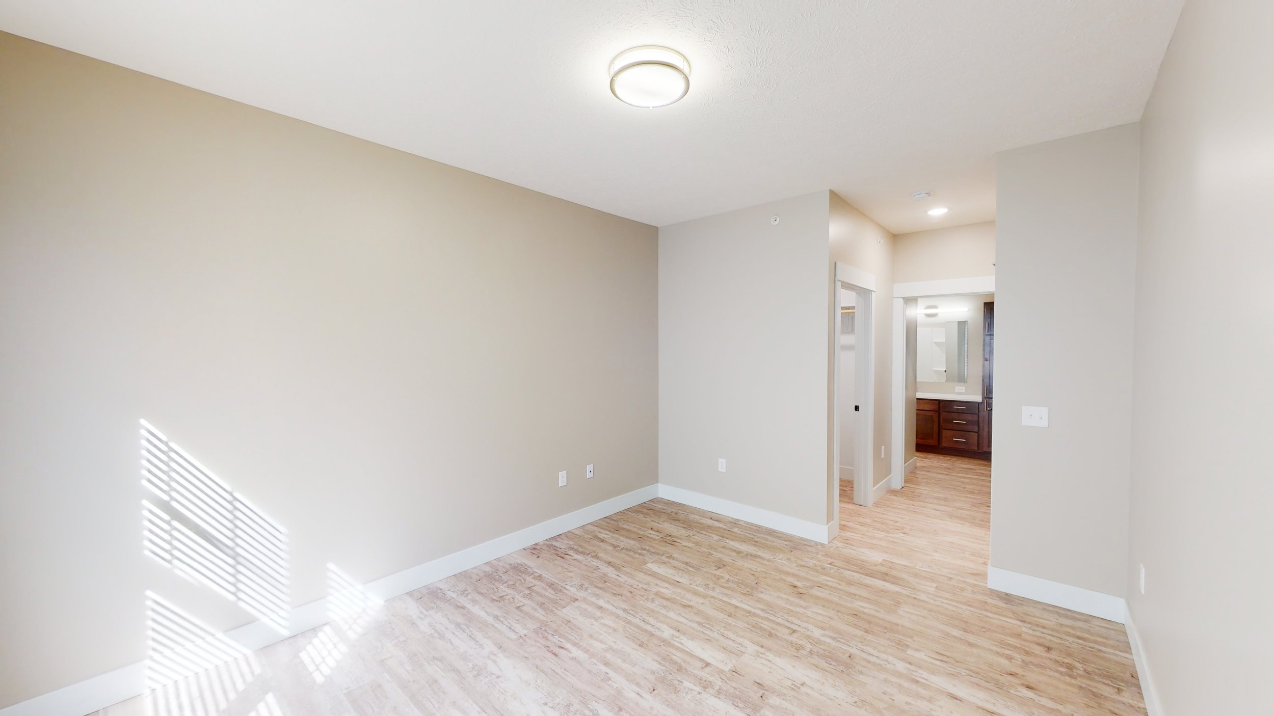 Empty room with light beige walls, wood flooring, ceiling light, and a doorway leading to a bathroom with a sink and mirror.