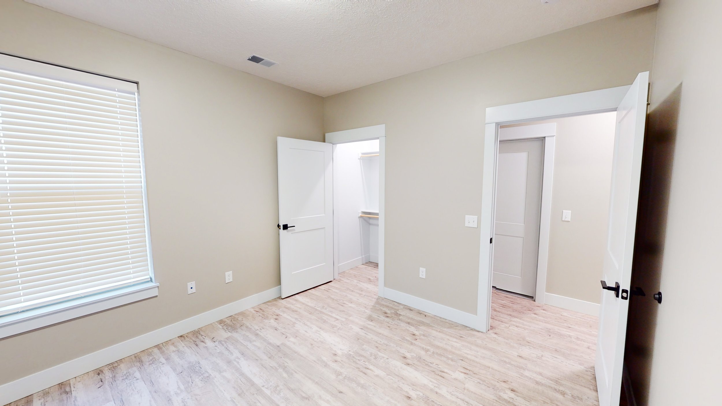 Empty room with wood flooring, light-colored walls, a window with blinds, and open doors to a closet and hallway.