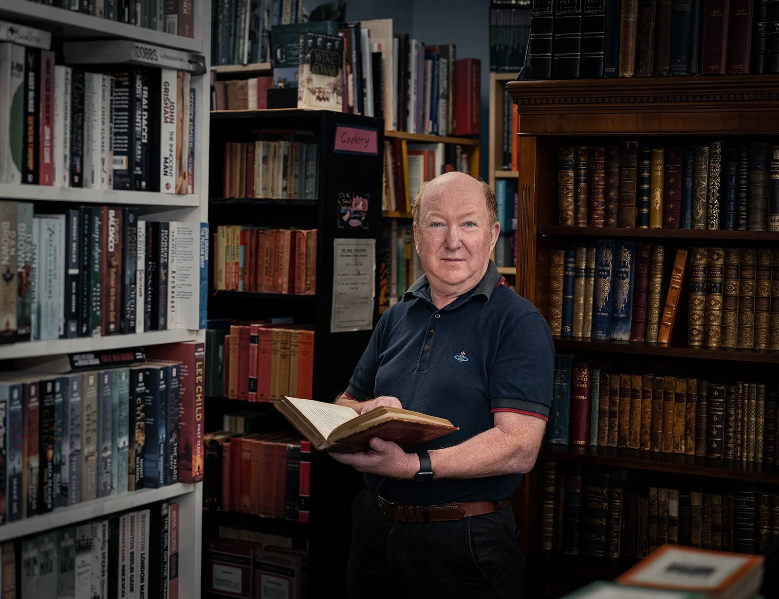 A man with light skin, wearing a dark polo shirt, stands in a bookstore or library holding an open book, surrounded by shelves of books.