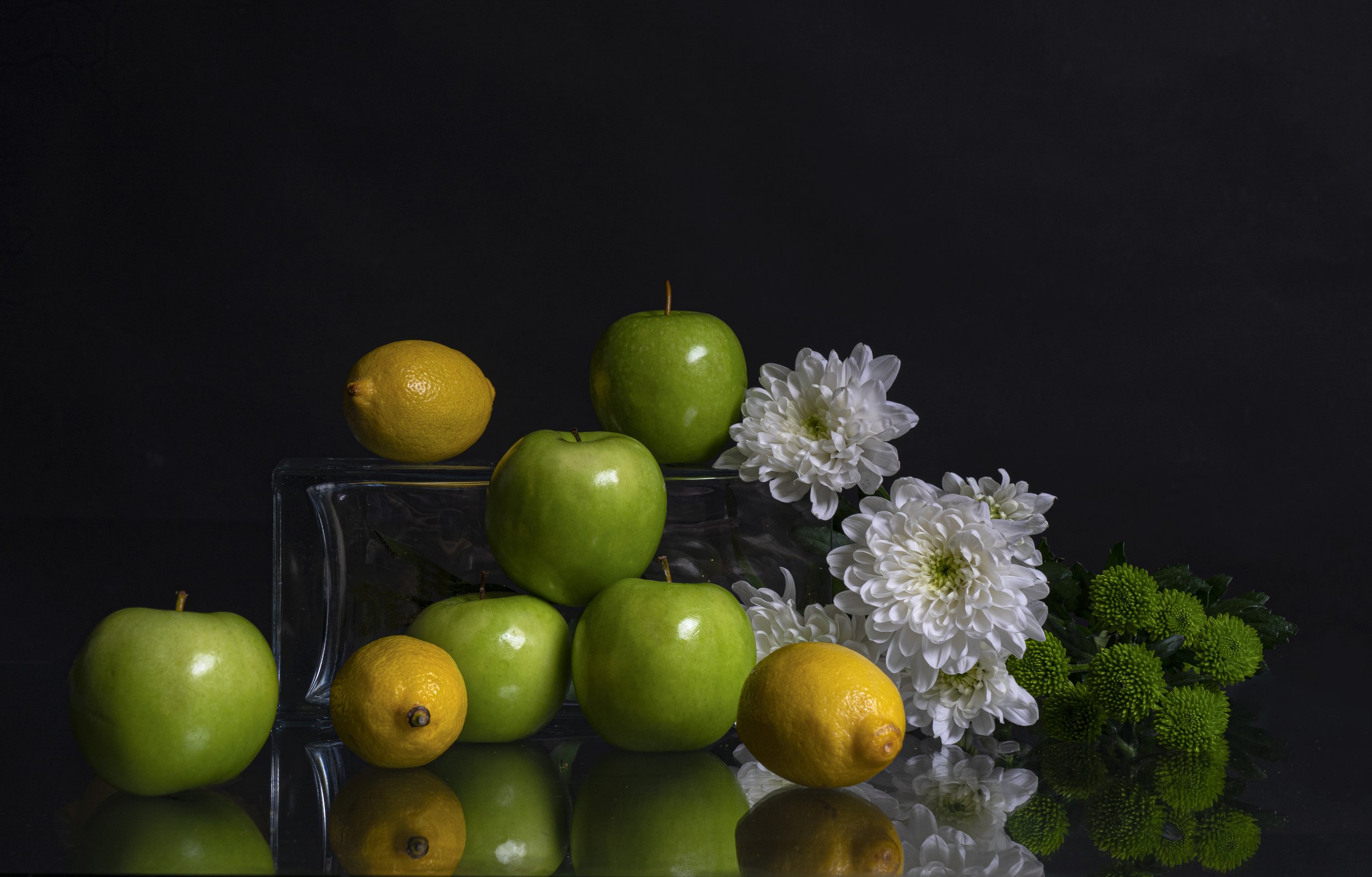 Green apples, yellow lemons, and white flowers on a reflective surface against a dark background.