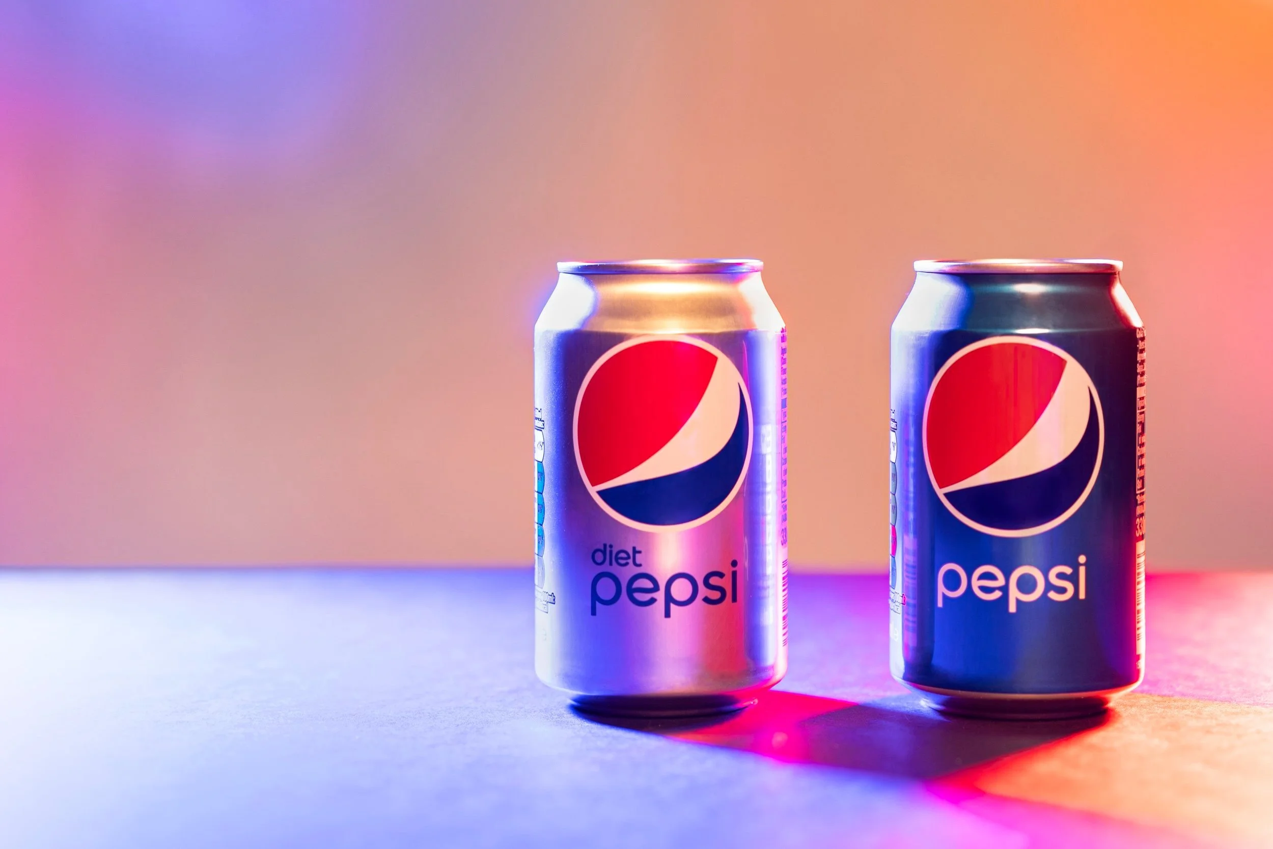 Two cans of Pepsi, one labeled Diet Pepsi, are positioned on a surface with colorful lighting creating a vibrant background and shadows.