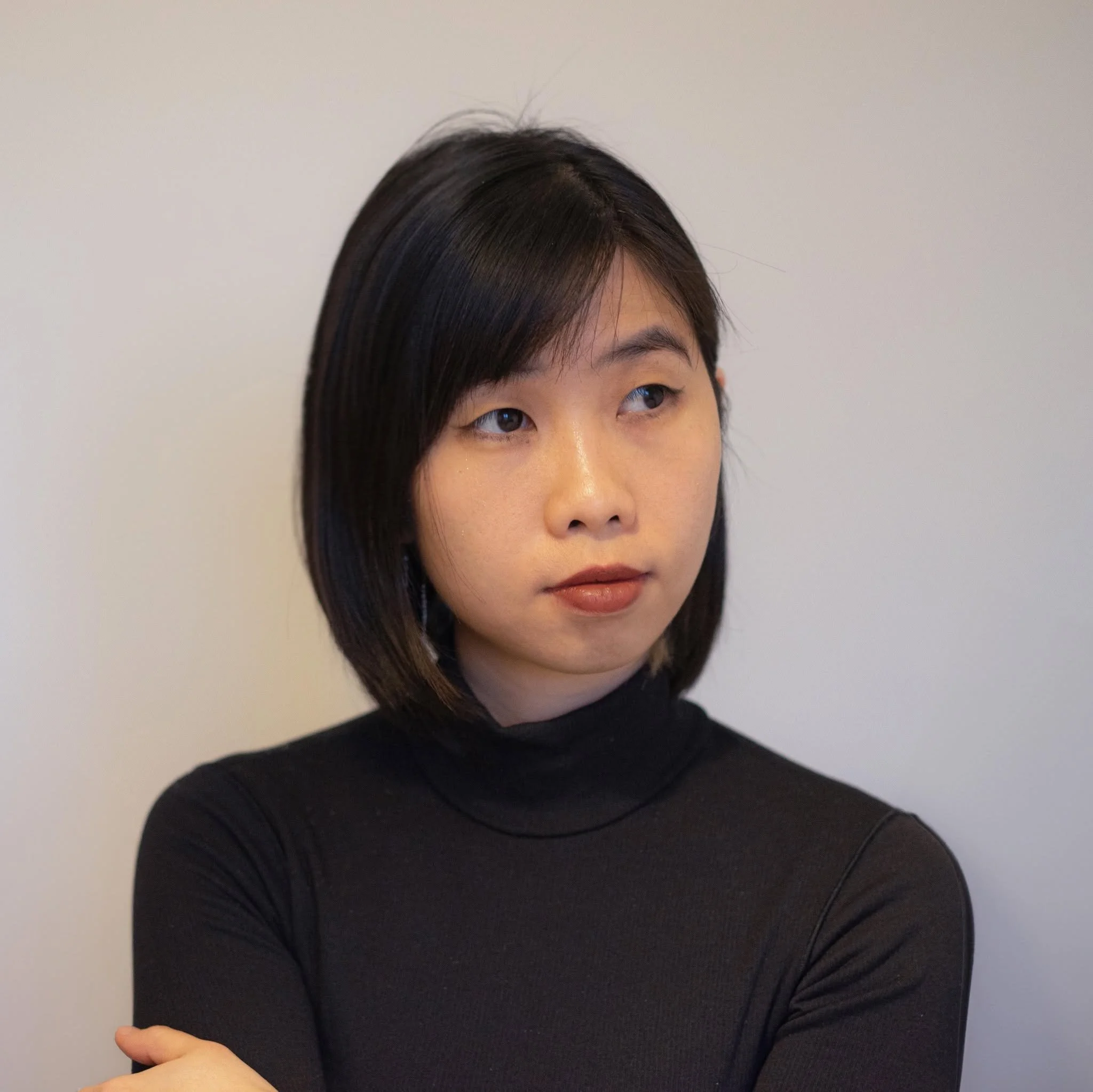 A person with short, dark hair wearing a black turtleneck shirt, standing against a plain light-colored background.