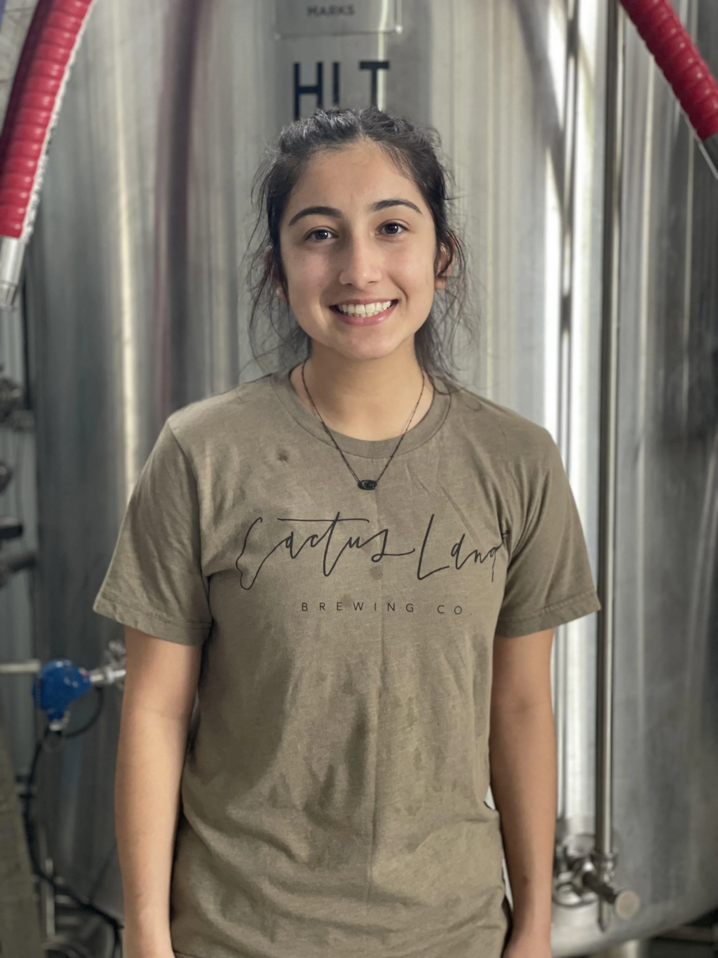 Person in front of brewing equipment wearing a "Cactus Land Brewing Co" t-shirt, smiling.