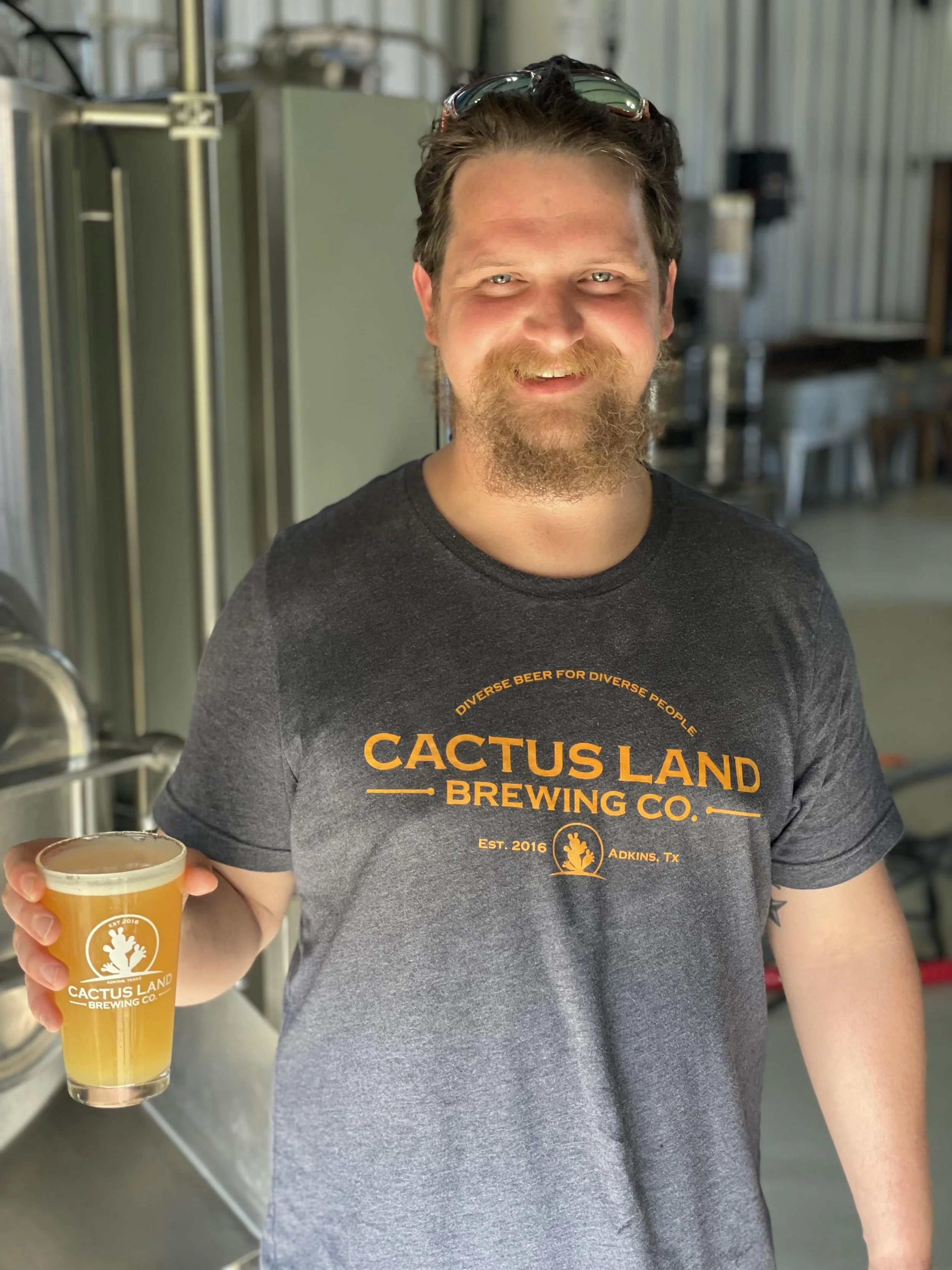 Man smiling while holding a glass of beer with brewing equipment in the background, wearing a Cactus Land Brewing Co. t-shirt.
