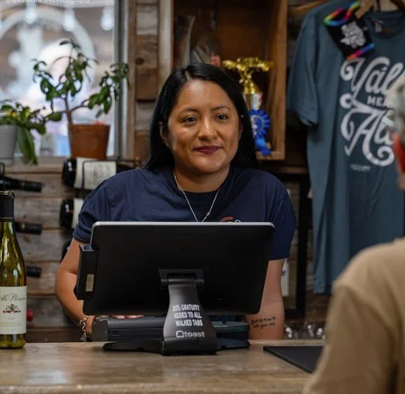 A woman standing at a register in a store, with a cash register and wine bottles visible, wearing a blue shirt, with a background of clothing and decor items.