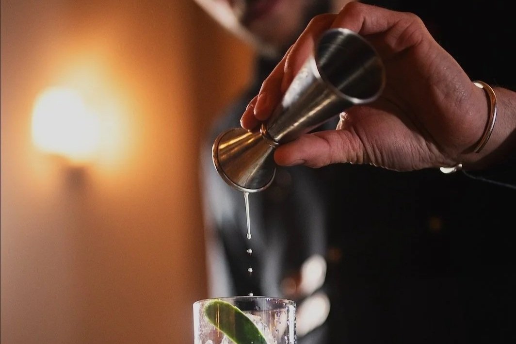 A person adding a liquid from a jigger into a glass with lime wedge.