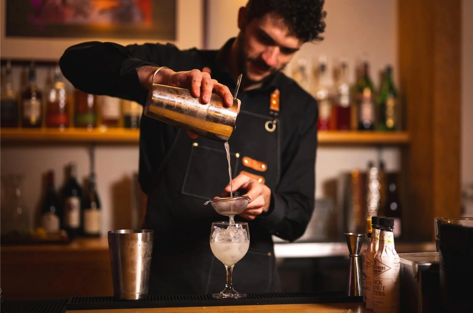 A bartender preparing a cocktail, pouring liquid through a strainer into a glass with ice, in a bar setting with bottles in the background.