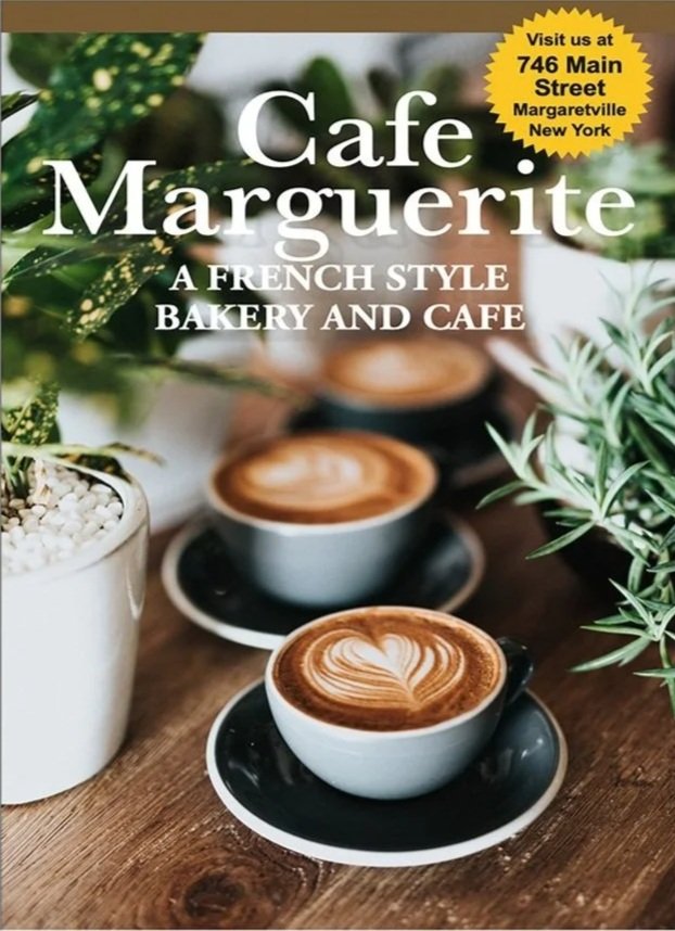 Three cups of coffee with latte art on a wooden table, surrounded by green plants, with a yellow sticker that says "Visit us at 746 Main Street, Margaretville, New York".