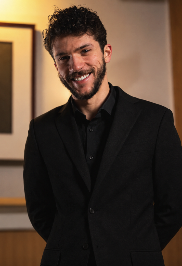 A smiling man with curly dark hair, beard, wearing a black suit and shirt, standing indoors with a blurred background of frames and warm lighting.