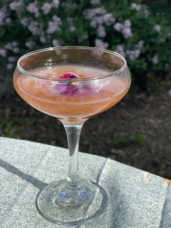 A pink cocktail garnished with an edible flower in a coupe glass on a patterned tablecloth outdoors, with greenery in the background.