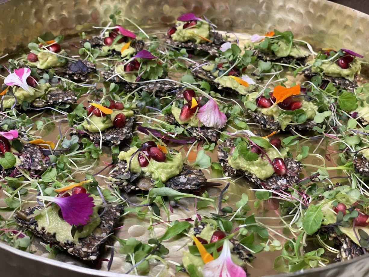 A tray of gourmet appetizers with seed-covered crackers topped with guacamole, pomegranate seeds, microgreens, and edible flower petals.