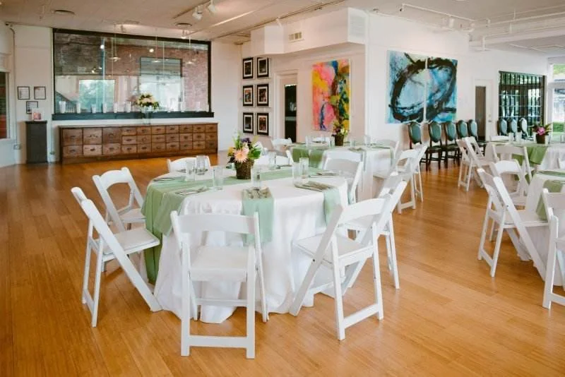Elegant event space with round tables set with white tablecloths and green runners, adorned with floral centerpieces, surrounded by white folding chairs; colorful abstract paintings decorate the walls.