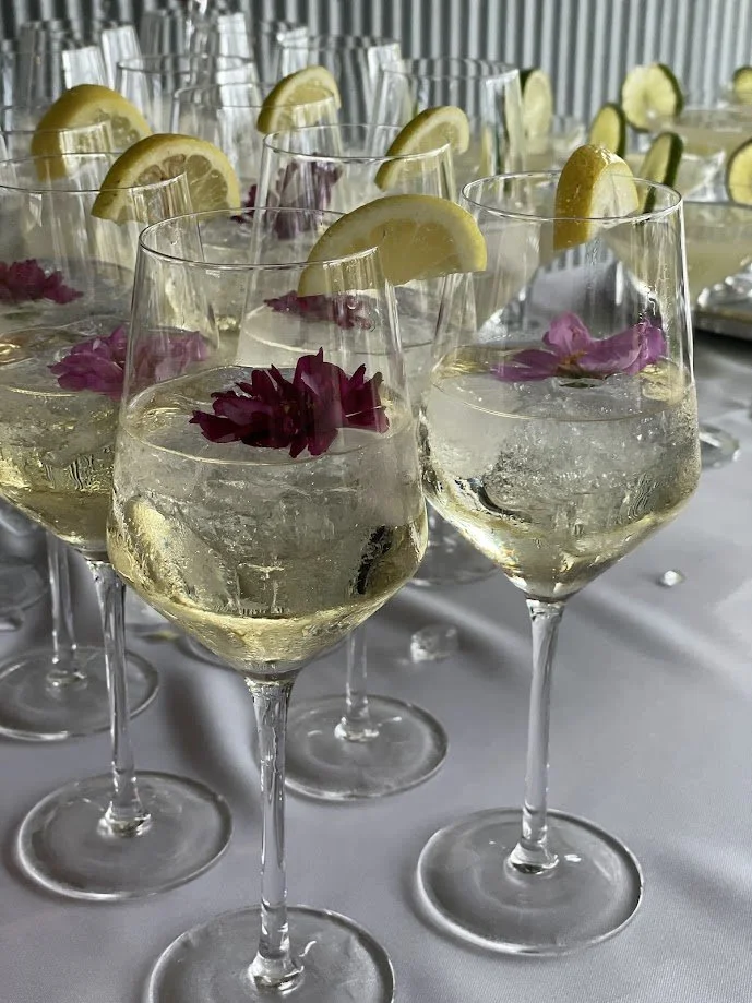 Multiple glasses of sparkling wine garnished with lemon wedges and edible flowers on a table.