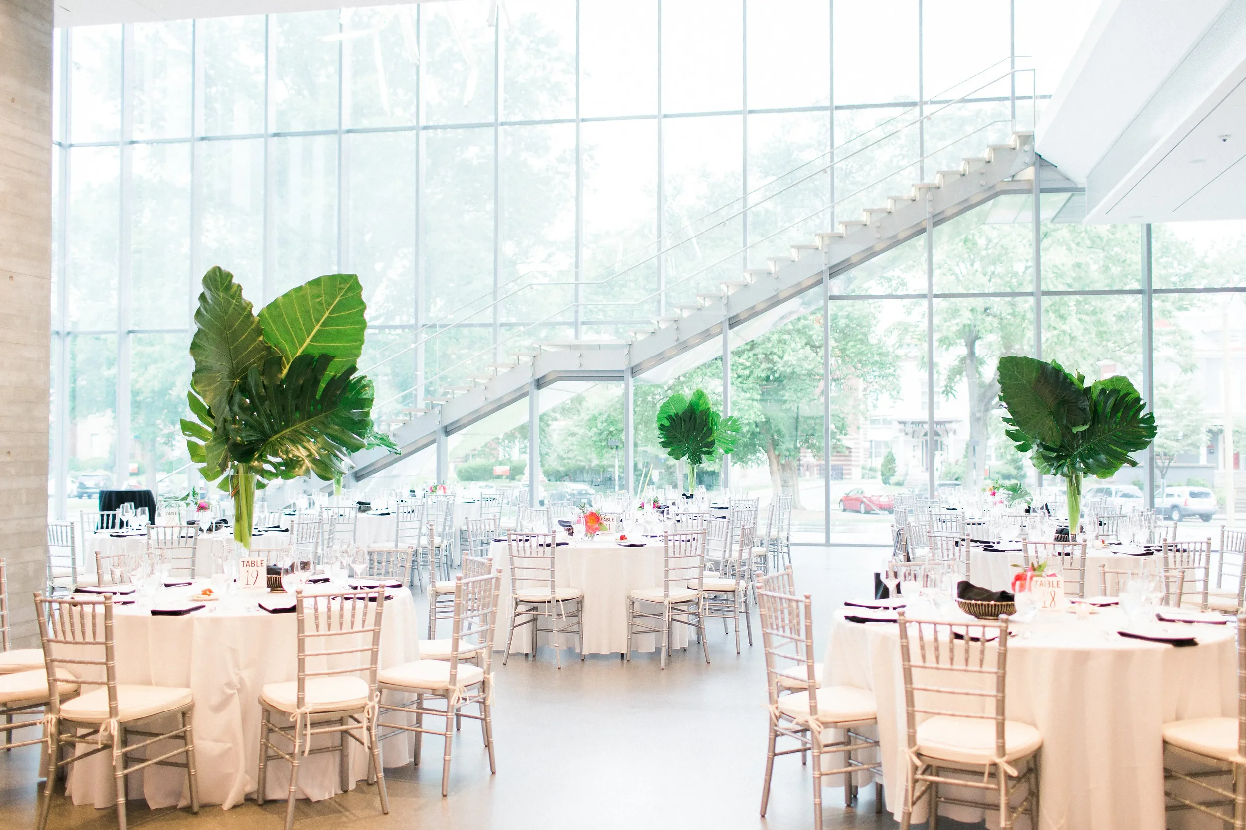 Interior of a modern event hall with round tables covered in white tablecloths, decorated with small centerpieces, and surrounded by white chairs. Large windows allow natural light to fill the space, and decorative plants are placed throughout the room.
