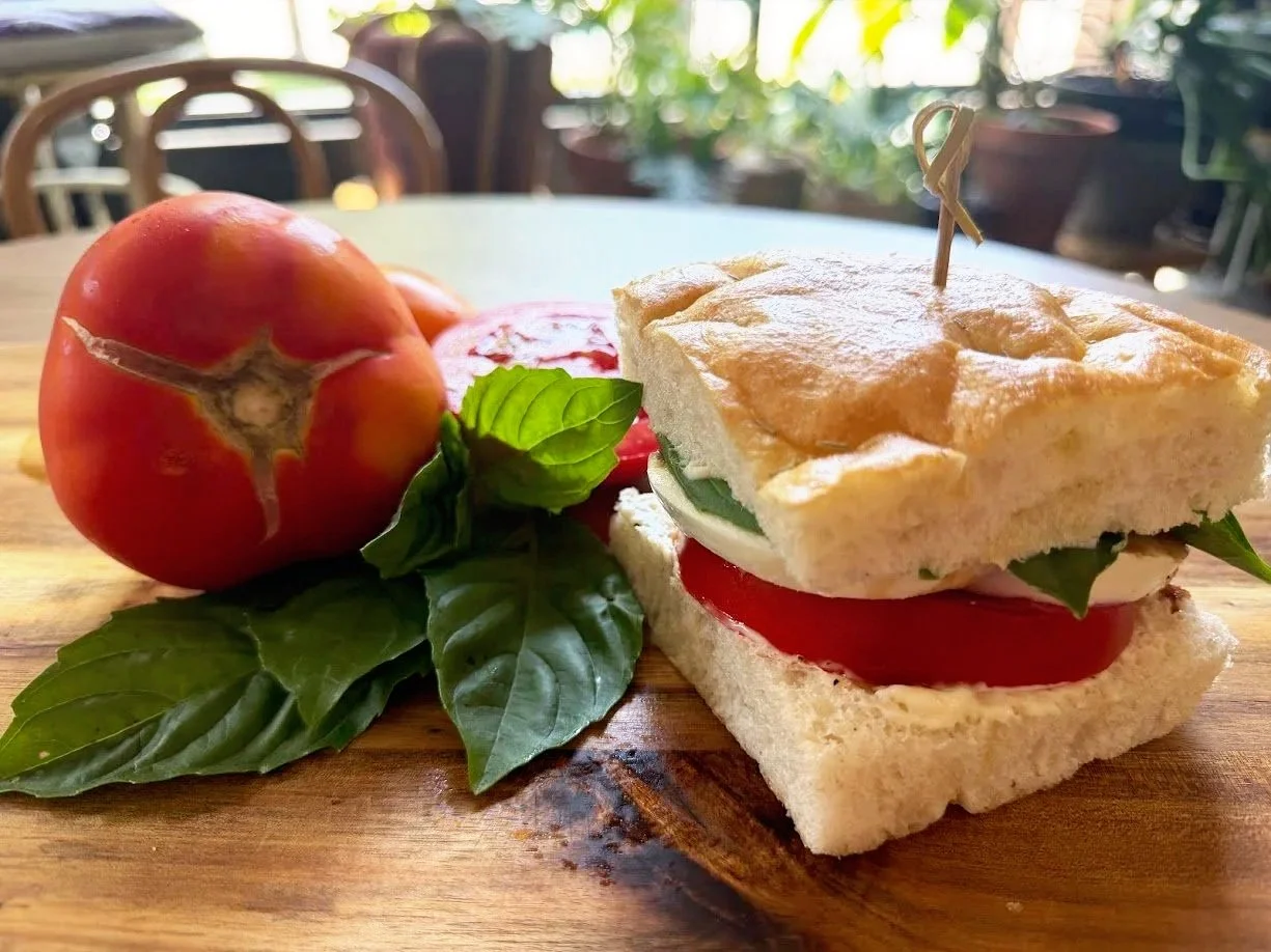 A sandwich with tomato, basil, and mozzarella on white bread, next to a whole tomato, on a wooden table in a bright outdoor setting.