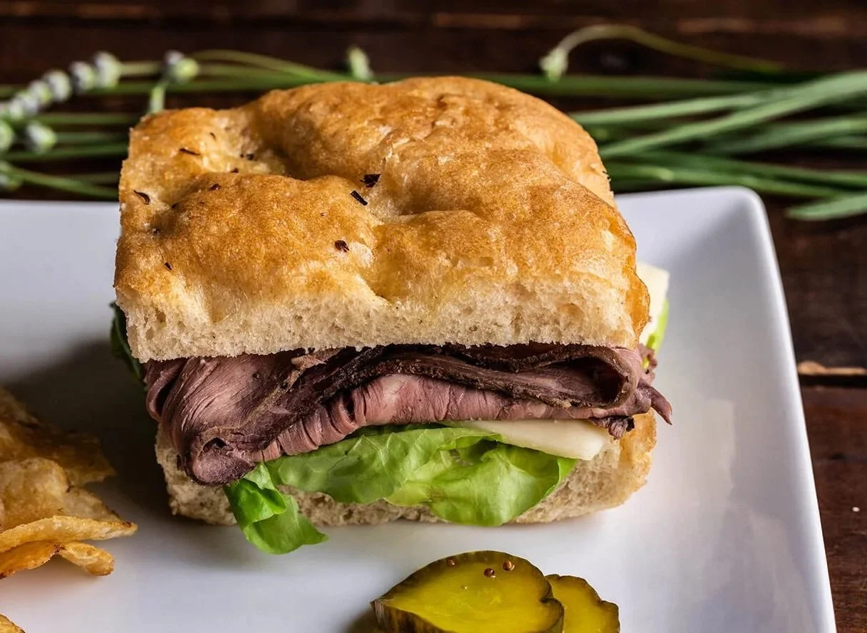 Beef sandwich with lettuce on a white plate, garnished with pickles, with potato chips nearby.