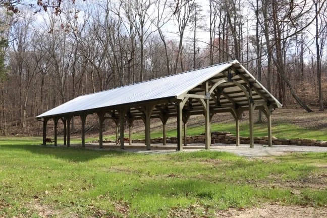 Open-sided wooden pavilion with a metal roof in a grassy area, surrounded by a forest of leafless trees.