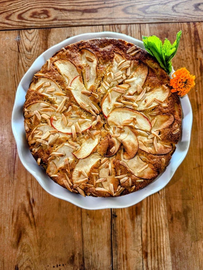 Apple cake topped with sliced apples and almond slices, garnished with fresh mint and an orange marigold flower in a white bowl on a wooden table.