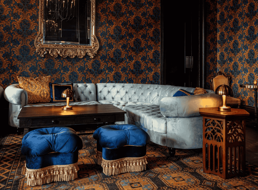 Luxurious living room with ornate wallpaper, a tufted sectional couch, blue velvet stools with fringe, a wooden coffee table, and gold accents.