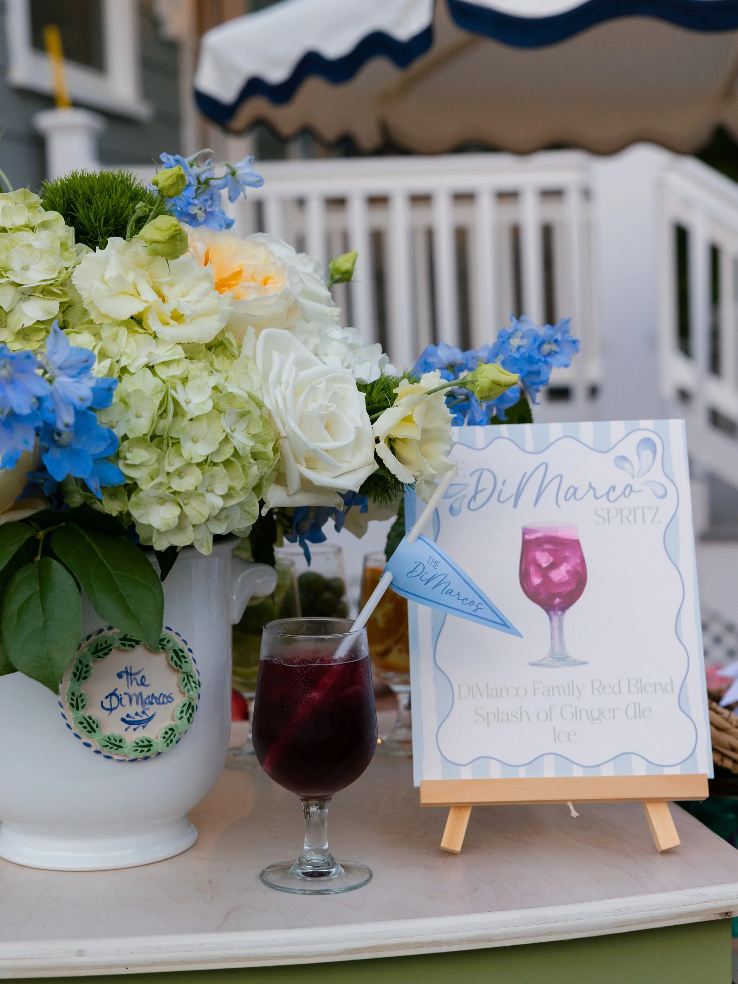 A floral arrangement with white roses, light green hydrangeas, and blue delphiniums on a table. There is a glass of beverage with a straw and a sign advertising a drink called 'DiMarco Spritz,' which is a red blend with ginger ale and ice. Photo by J