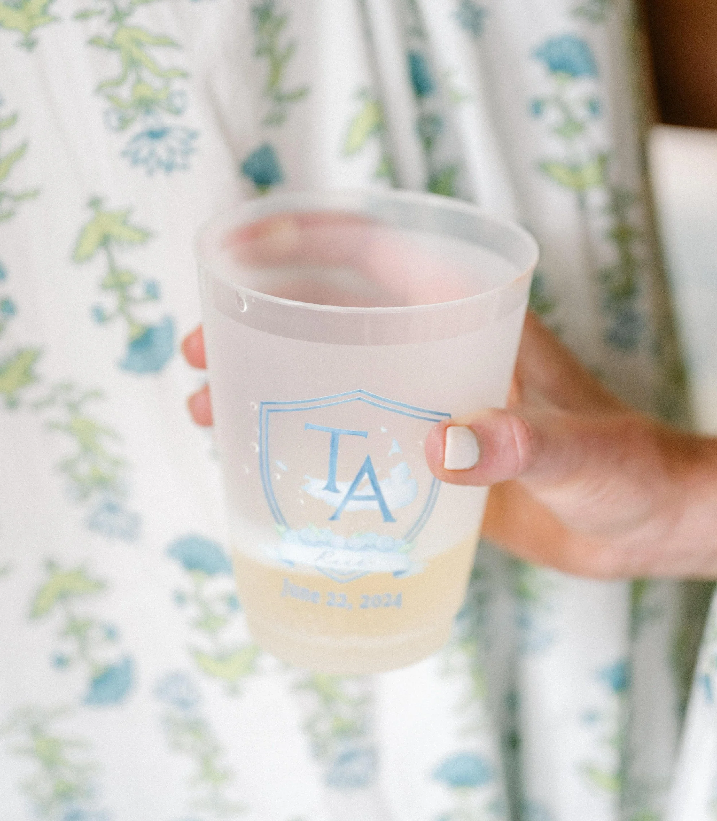 Person holding a plastic cup with a light-colored beverage, with a monogram and date printed on the cup, in front of someone wearing a floral dress.