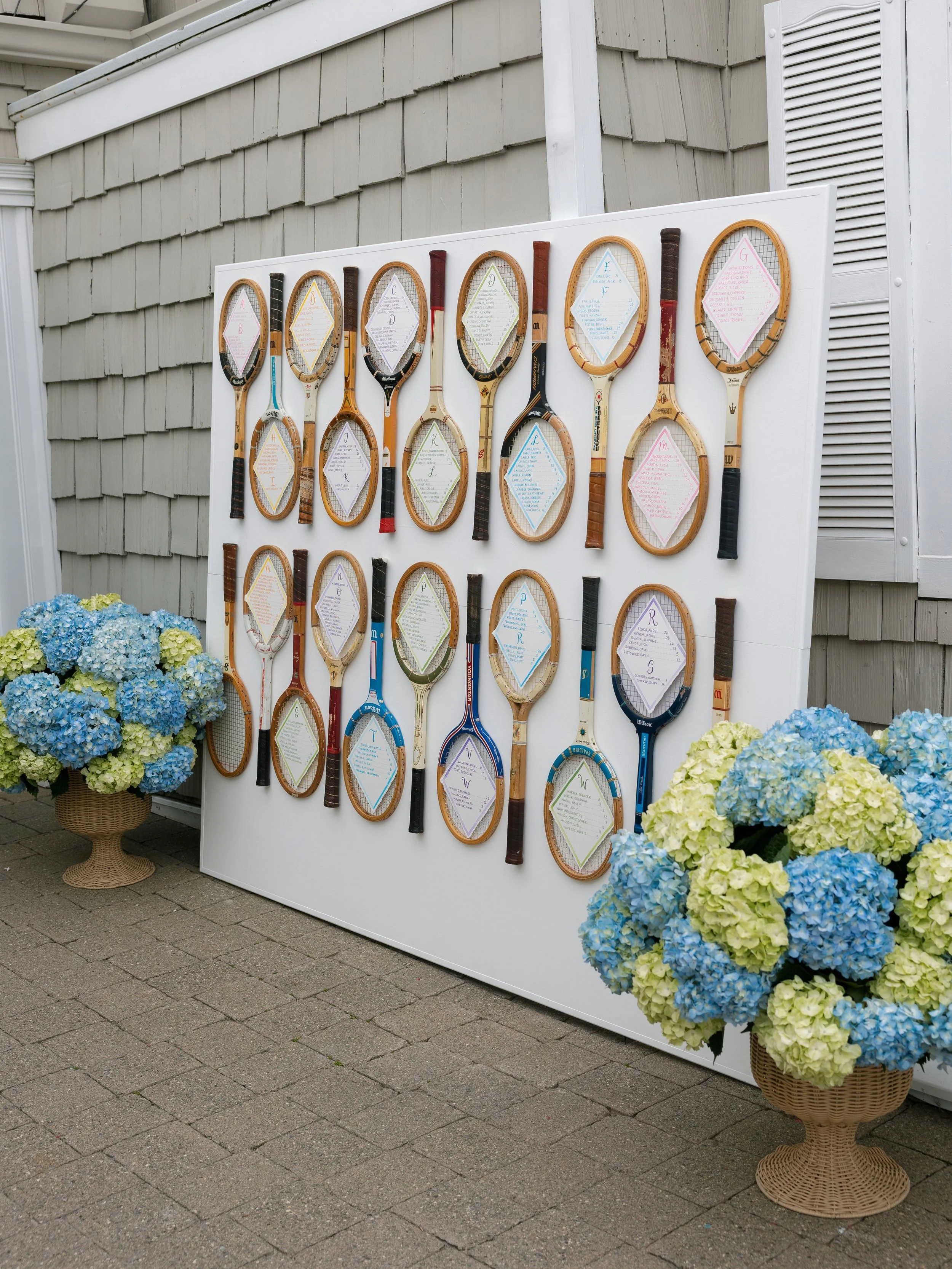 An outdoor display of vintage tennis rackets with paper notes inside their frames, arranged on a white board. Two large baskets of blue and green hydrangeas are placed on either side of the display. Photo by Jessa Schifilliti.