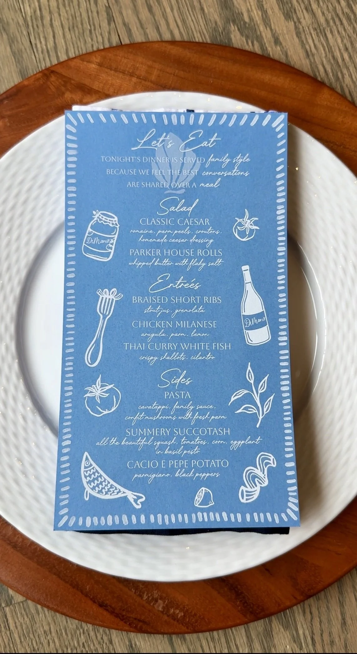 Blue dinner menu with handwritten-style text and white illustrations on a white plate, placed on a wooden table.