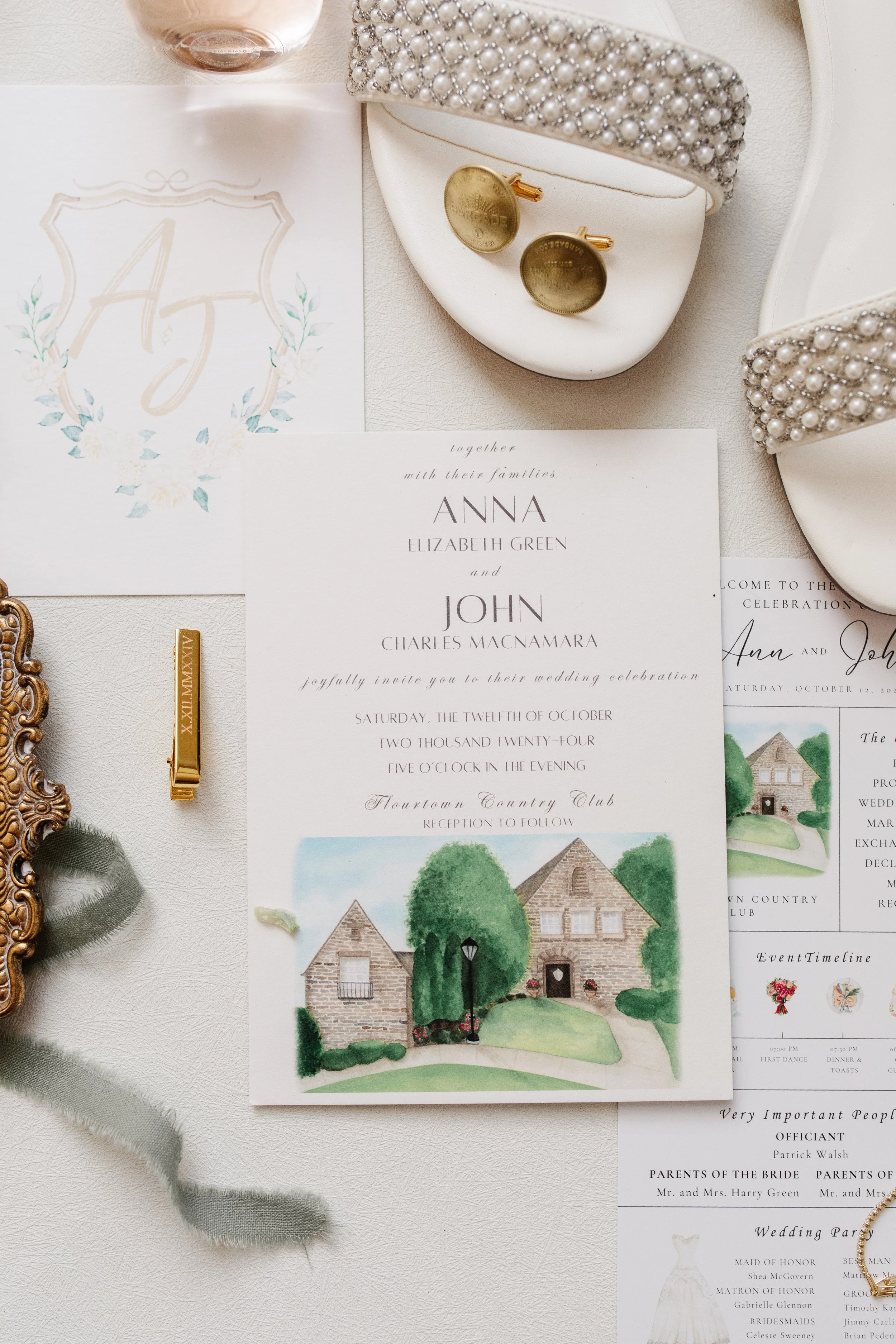 Wedding invitation card with watercolor illustration of a country house, surrounded by trees and a lamppost, placed on a white surface next to pearl-embellished shoes, gold cufflinks, and a decorative gold frame.