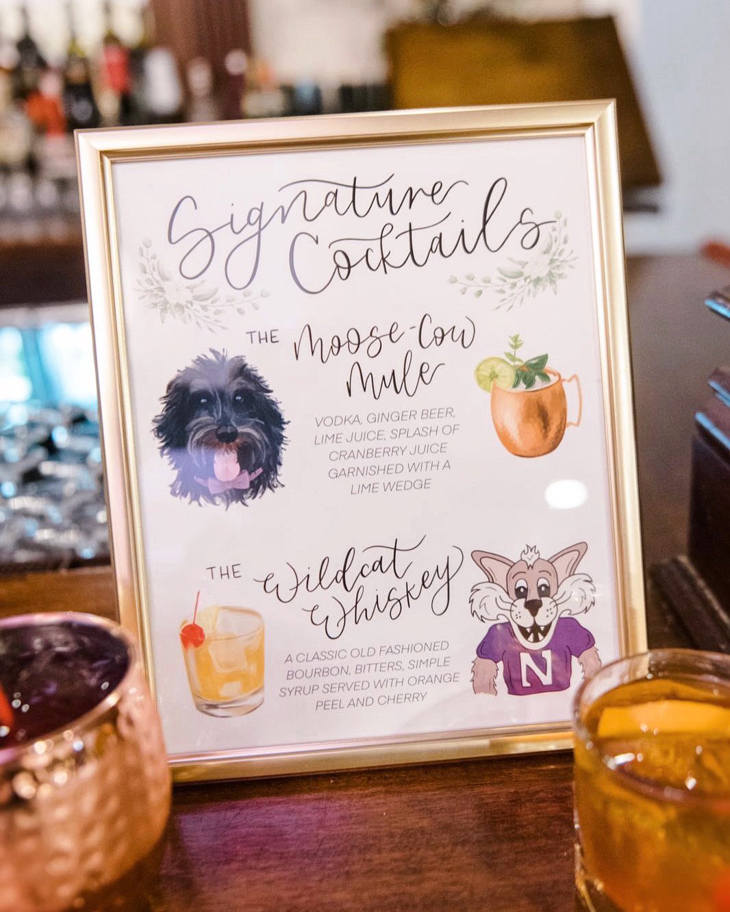 Menu sign with illustrations of a dog and a cartoon wolf, featuring signature cocktails named 'The Moose-Cow Mule' and 'The Wildcat Whiskey,' with drinks and garnishes shown.
