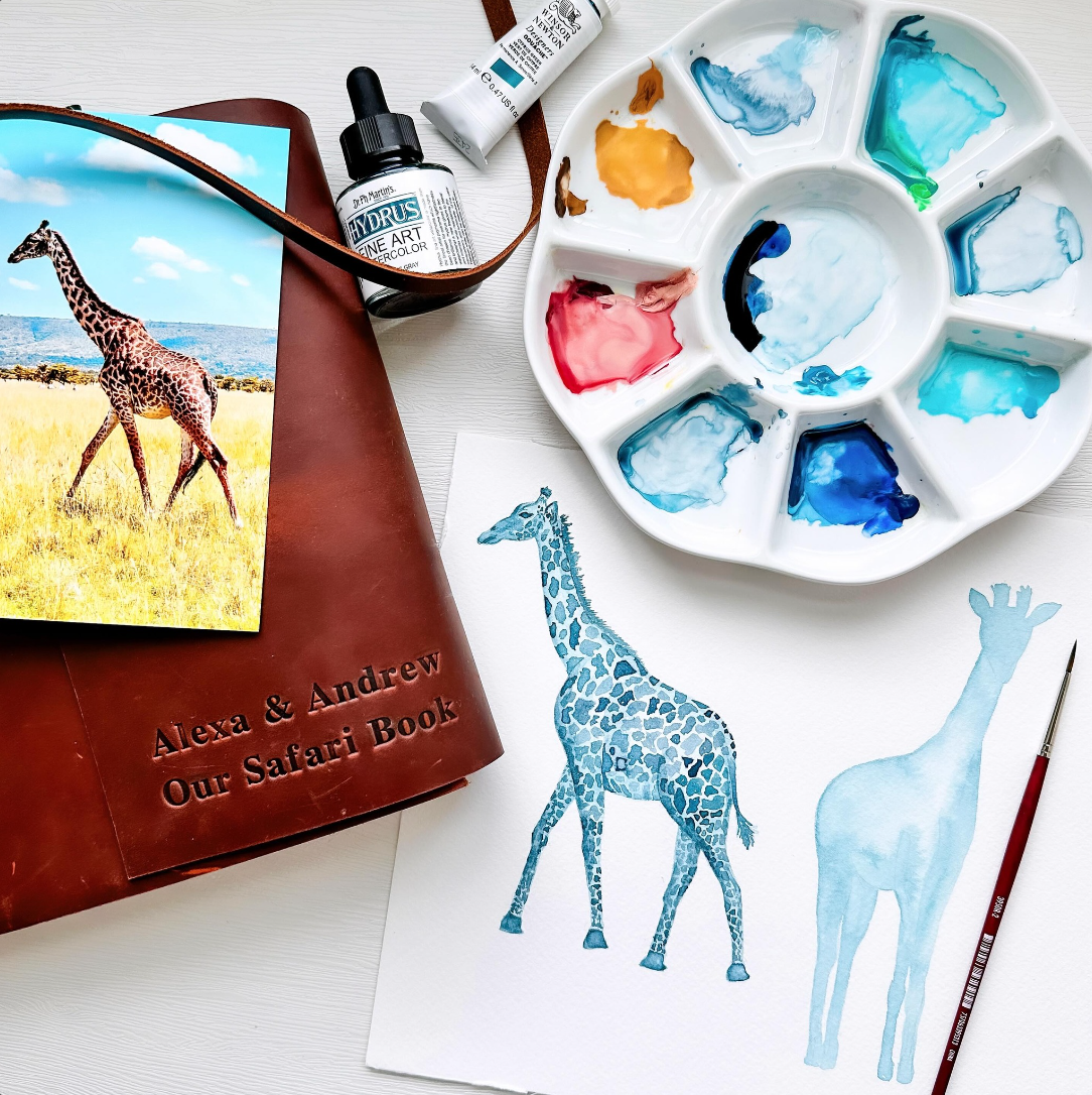 Watercolor painting supplies, including a palette with blue, yellow, and pink paint, a small watercolor brush, and two illustrations of a giraffe on paper. A photo of a giraffe in the wild is on a leather-bound book titled "Alexa & Andrew Our Safari 