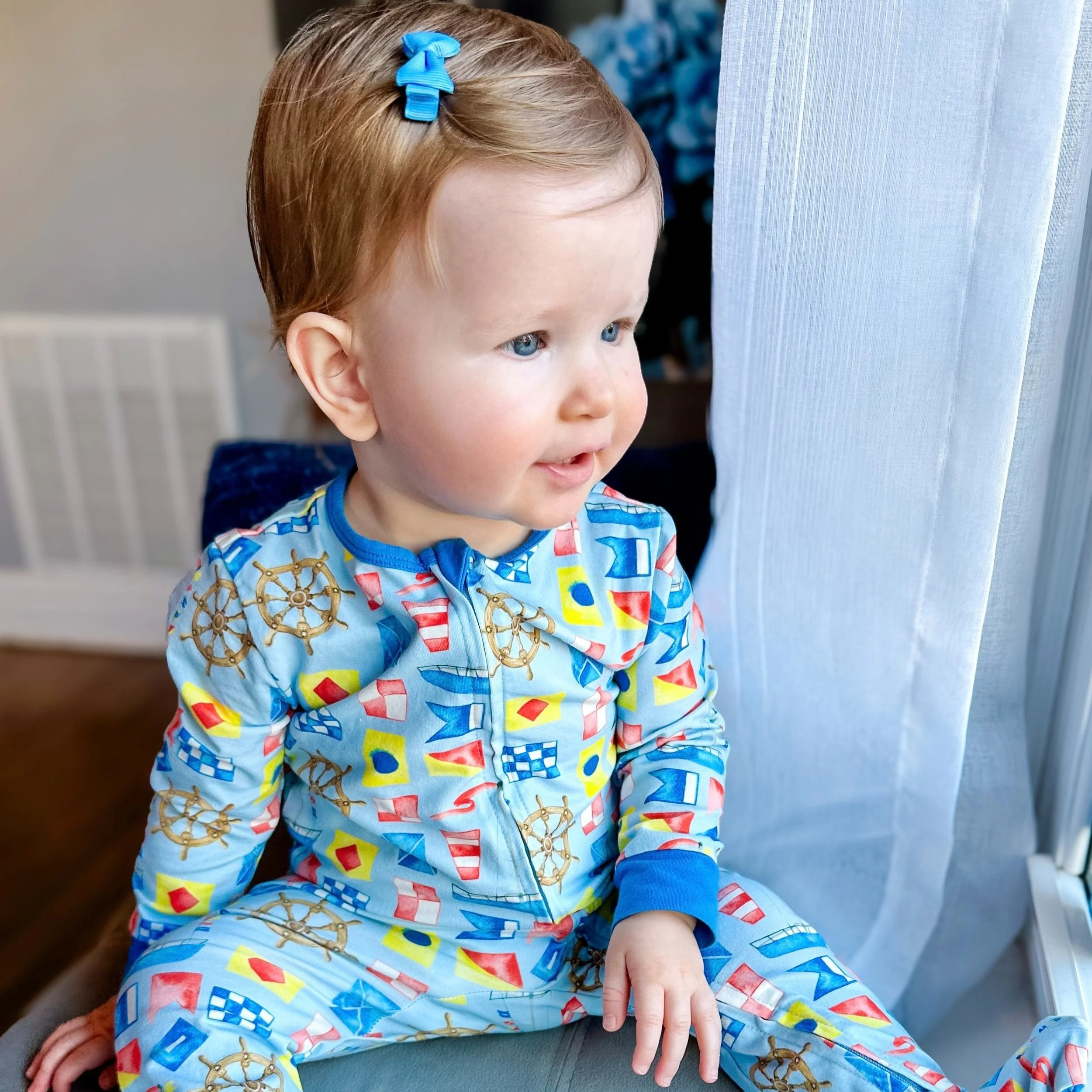 A young child with red hair and blue eyes sitting by a window, wearing a colorful nautical-themed outfit with ship wheels and geometric shapes, looking outside.
