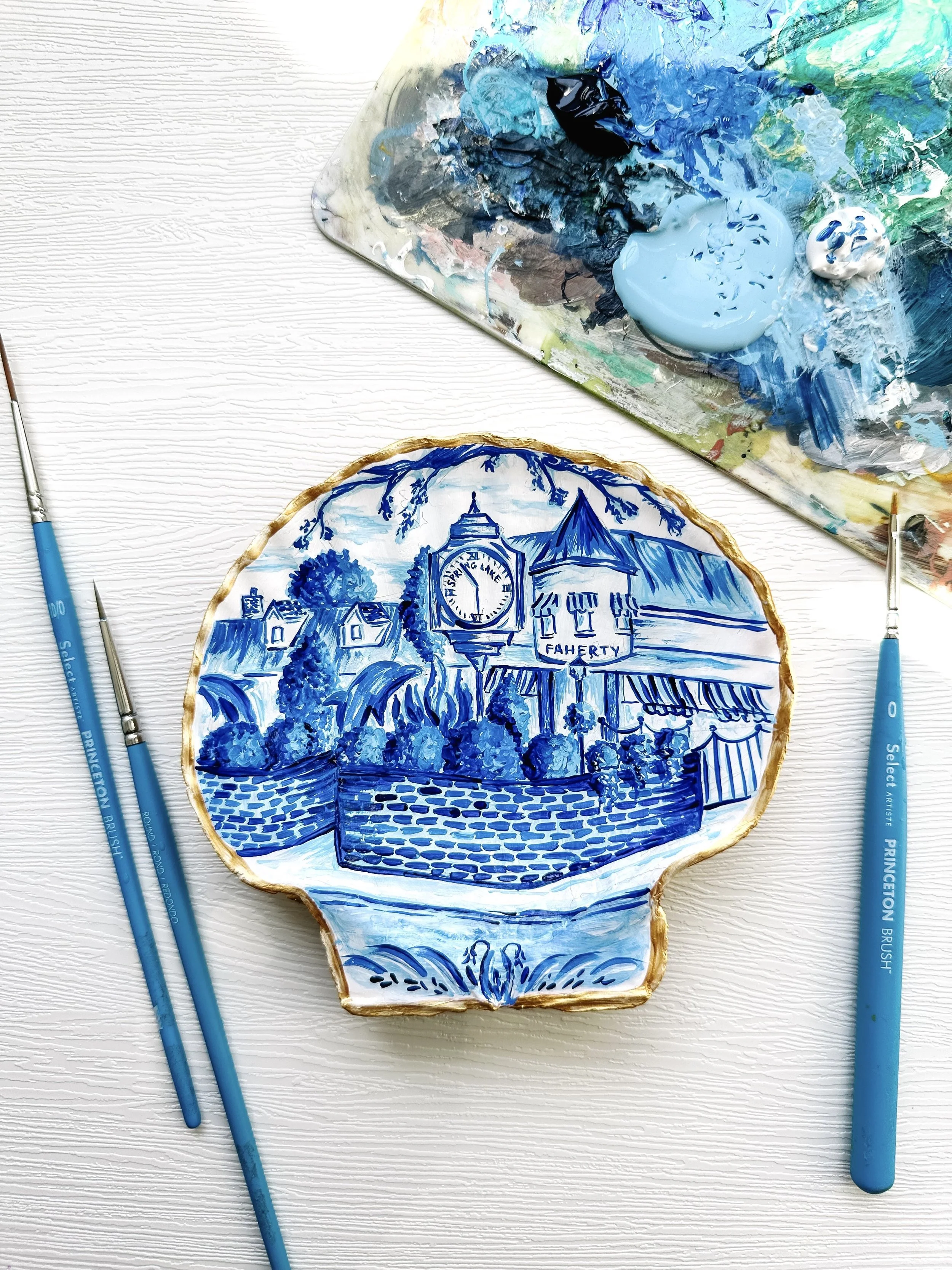 Decorative ceramic plate painted with blue and white landscape scene featuring a clock tower, buildings, and trees, placed on a white textured surface surrounded by two paintbrushes and a palette with blue and black paint.
