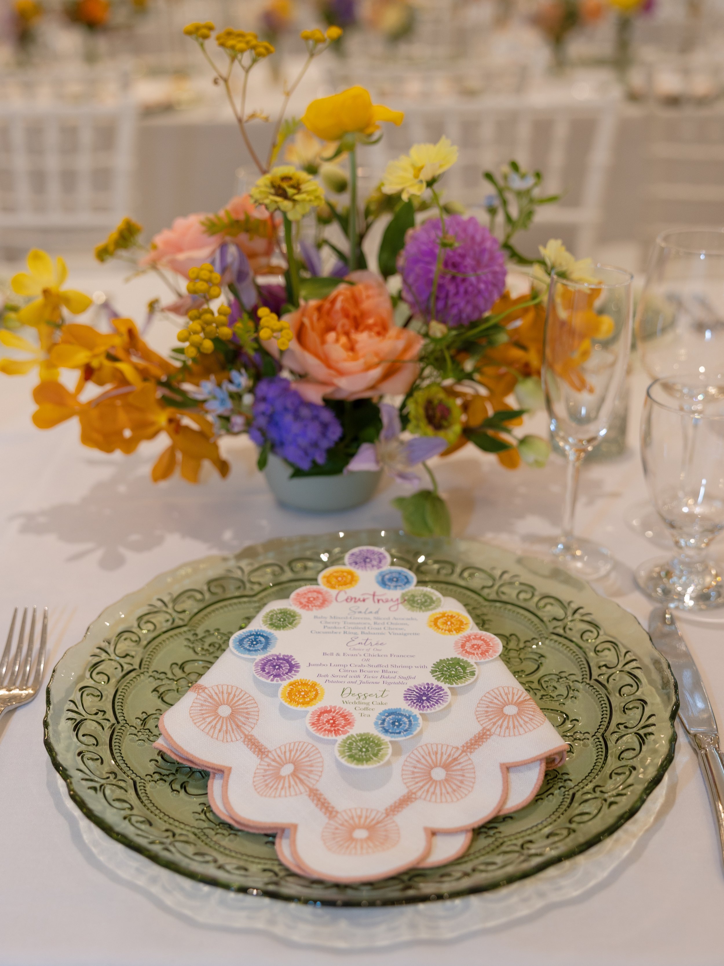 A table setting with a green glass plate, a floral napkin with a colorful menu, a centerpiece of multicolored flowers, and empty wine glasses. Photo by Jessa Schifilliti.