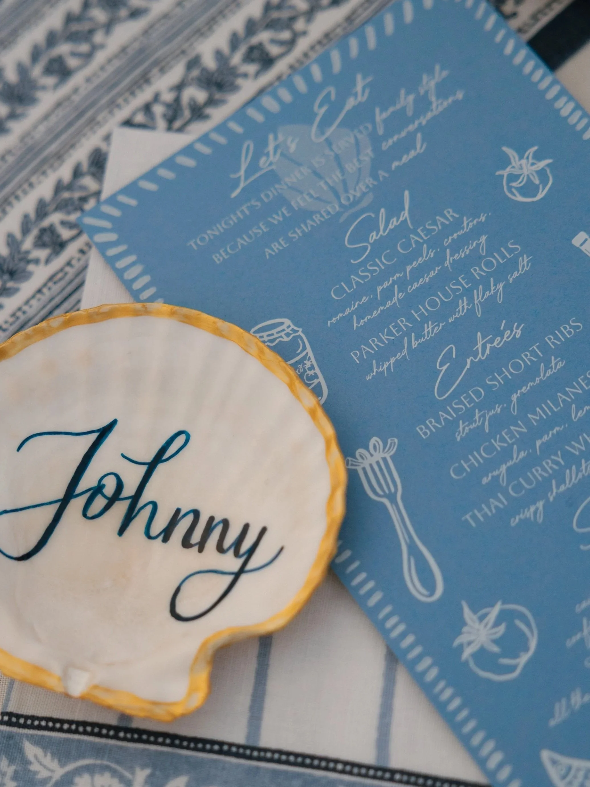 A blue menu with white text and illustrations, a white shell-shaped sign with the name Johnny in dark blue, and a tablecloth with a blue and white pattern. Photo by Jessa Schifilliti.