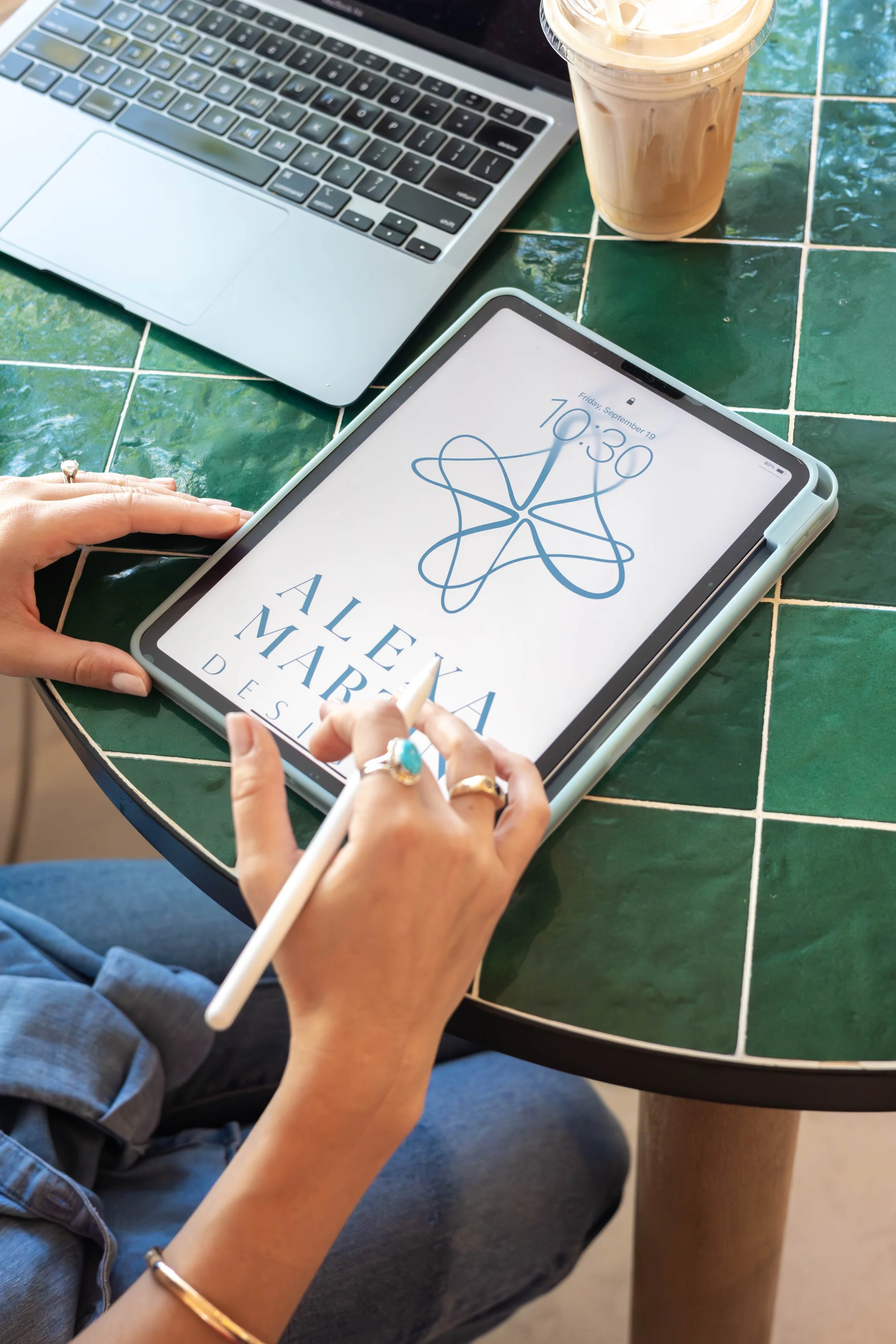 A person using a stylus on an iPad with a logo design and the text 'ALEXA MARA DESIGN', on a green-tiled table. There is a laptop and a iced coffee with cream in a plastic cup nearby.