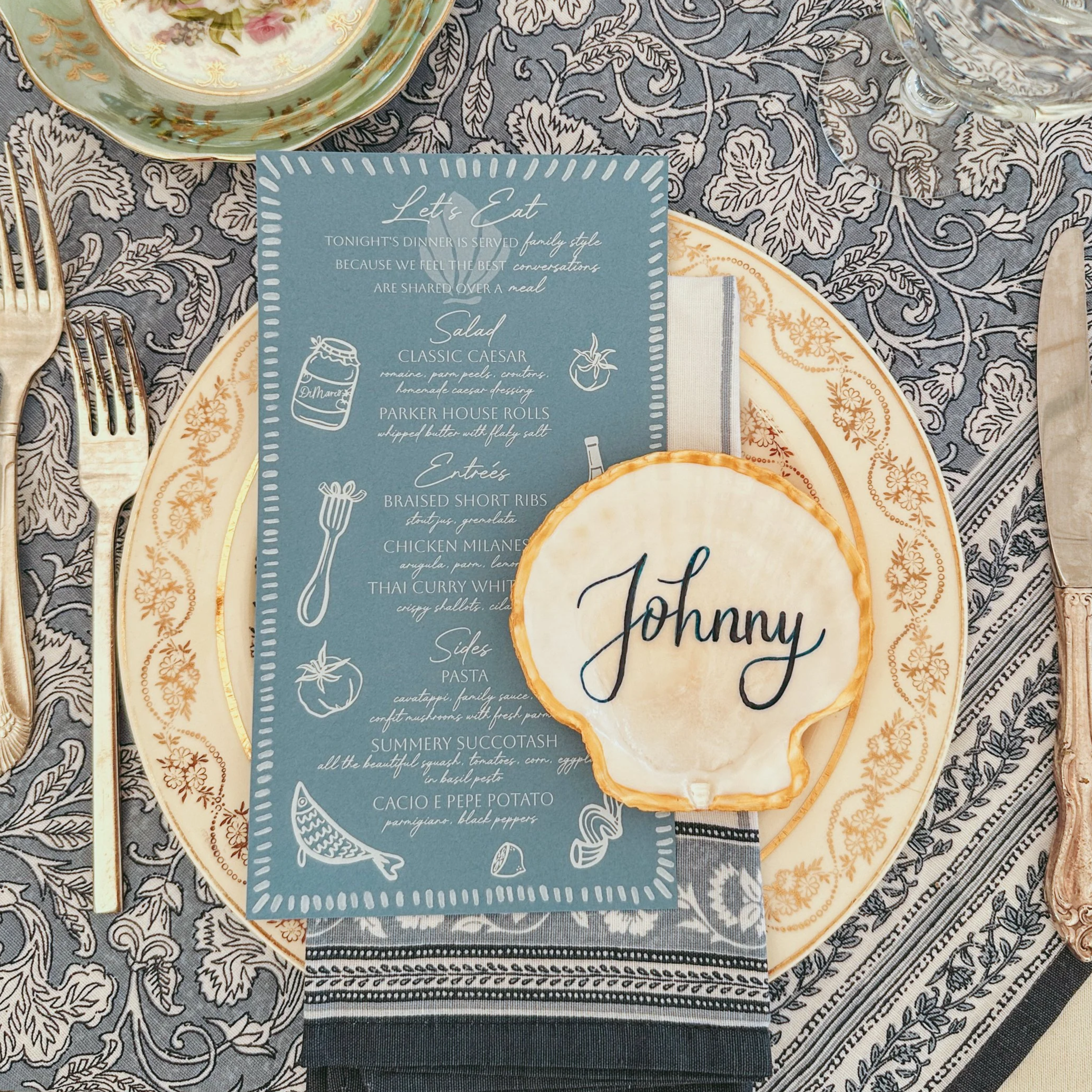 A dinner table setting with a decorative plate, silverware, a personalized cookie with 'Johnny' written on it, and a blue menu card listing a meal menu, on a patterned tablecloth.
