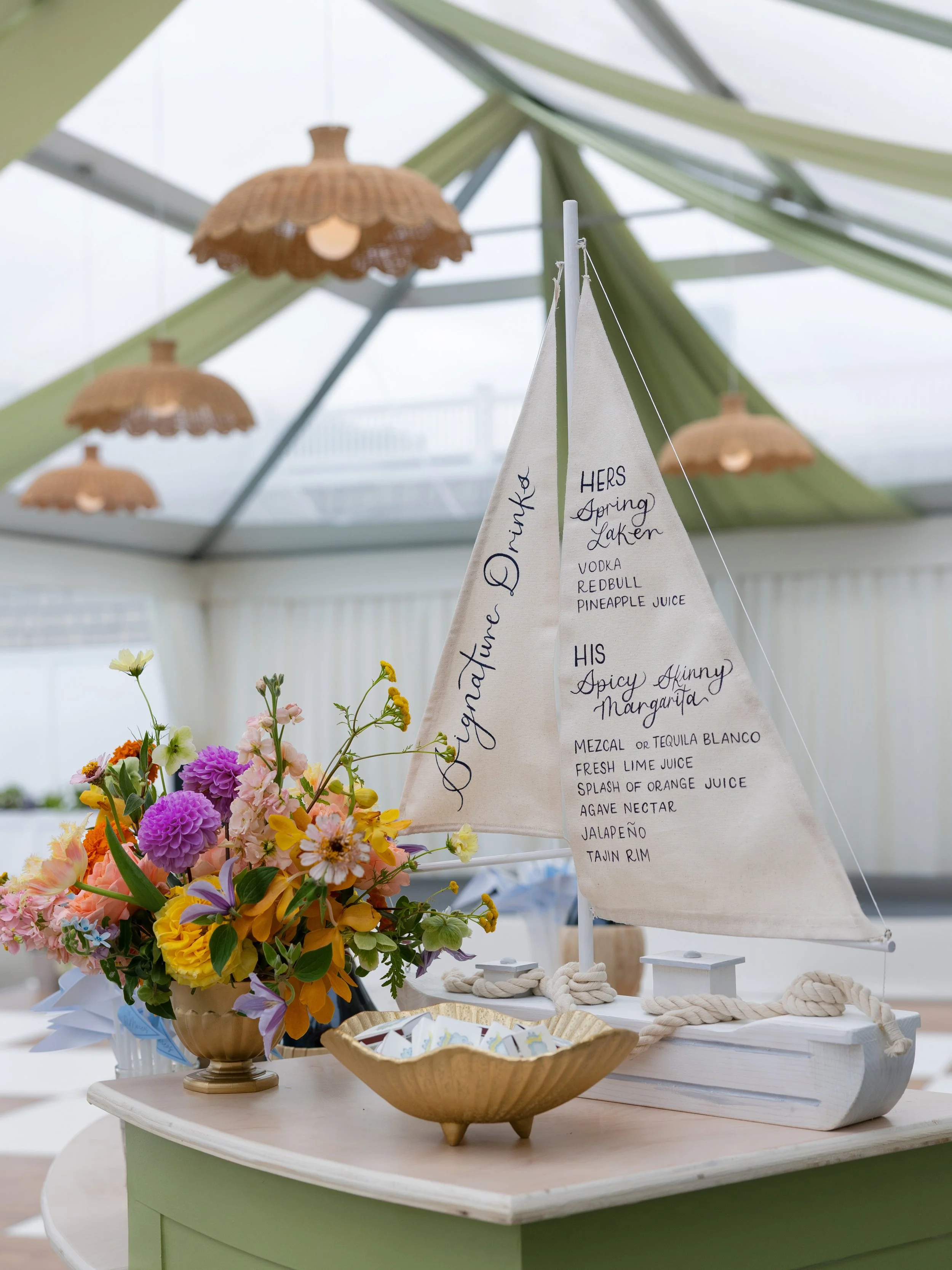 Decorative sailboat with beverage list, colorful flowers in a vase, and a rope on a white table inside a glass-ceiling room.