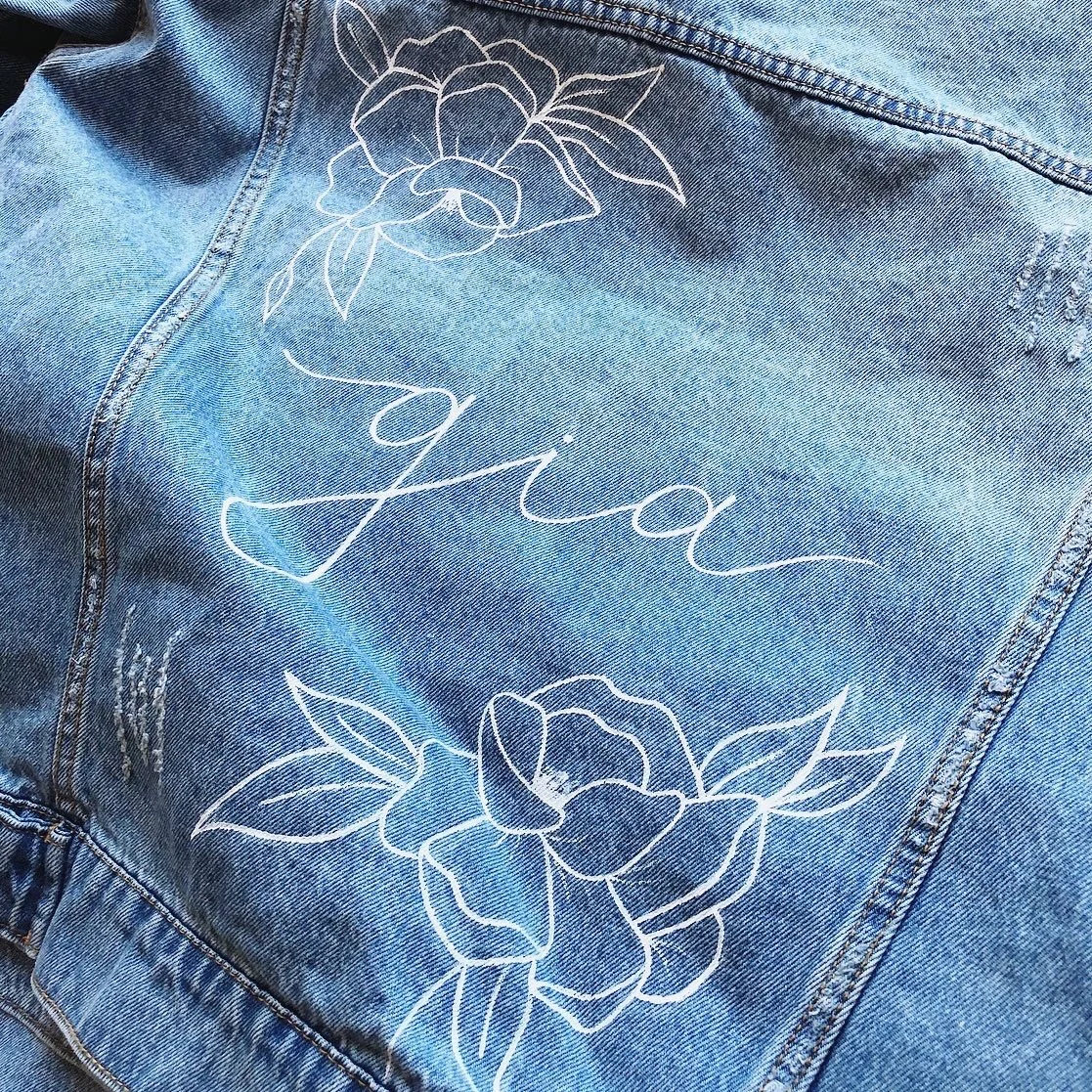 Back pocket of a blue denim jacket with white embroidered floral design and the word "giau" in cursive.