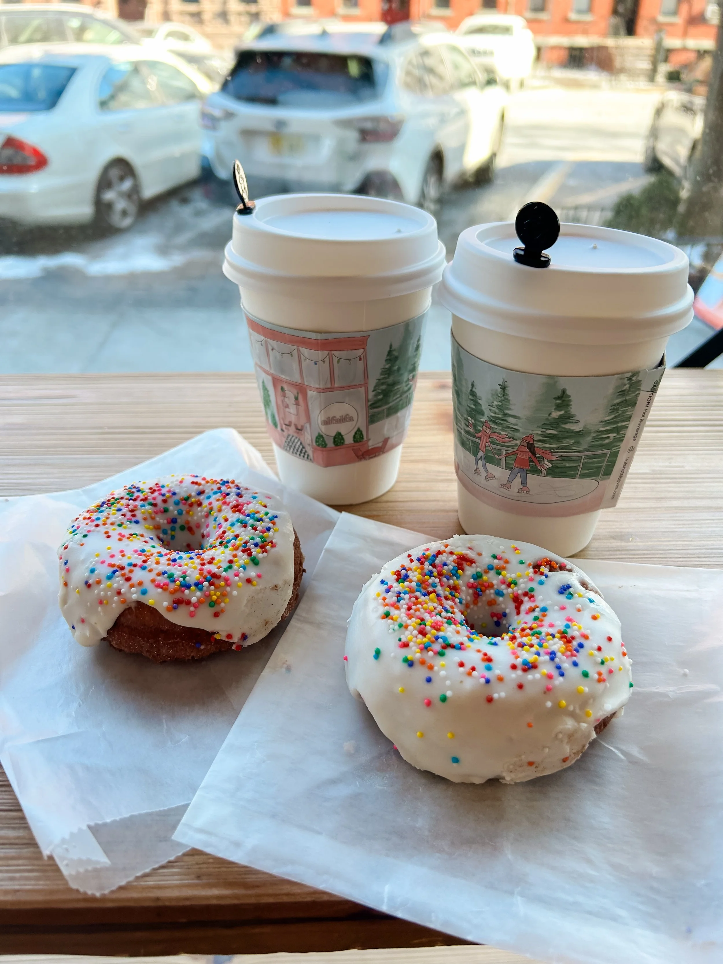 Two donuts with white icing and colorful sprinkles on white paper, two hot beverages in paper cups with holiday-themed sleeves, placed on a wooden table near a window with cars outside.