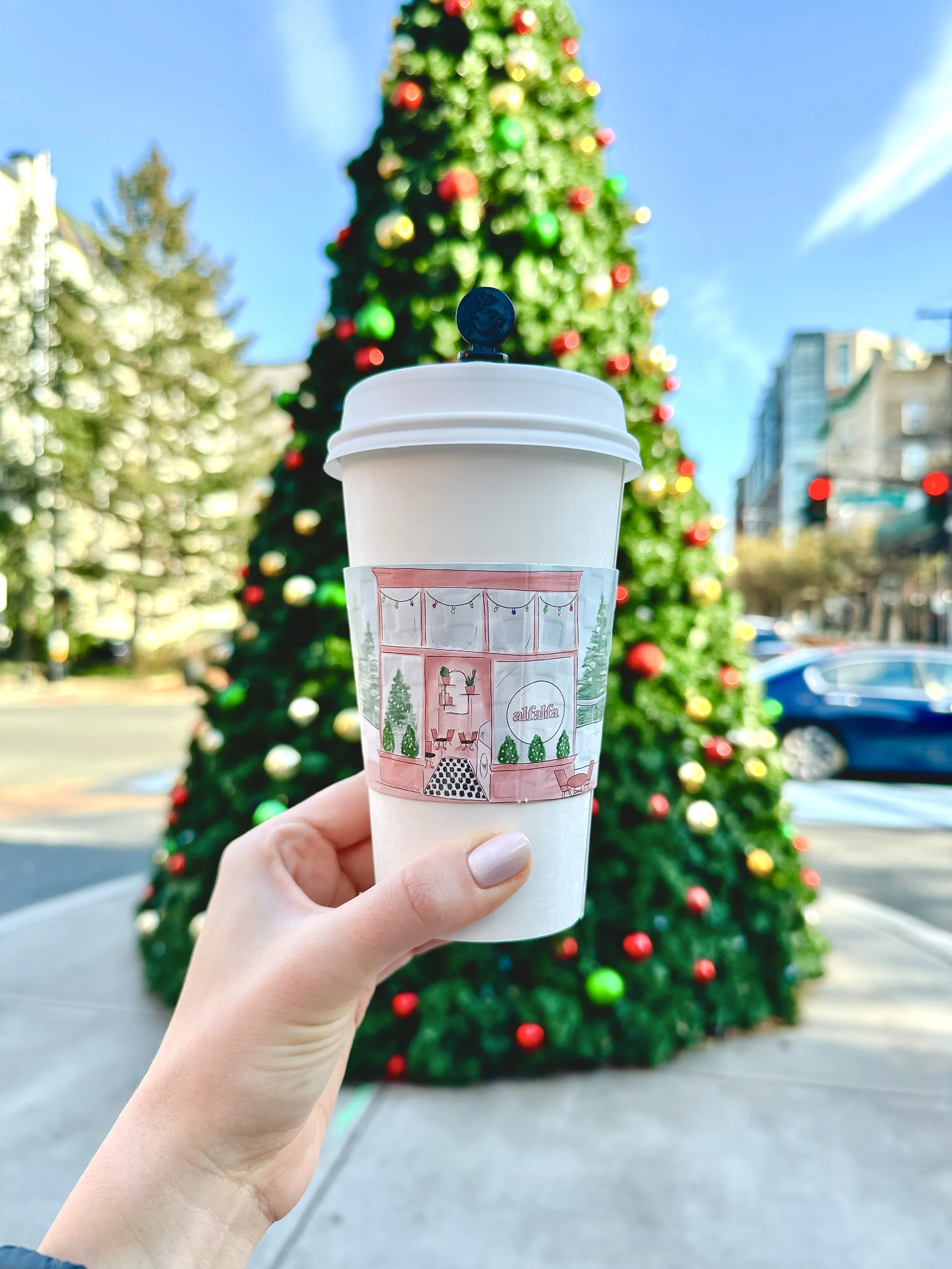 Person holding a to-go coffee cup with a drawing of a decorated Christmas interior, in front of a large, decorated Christmas tree outside during daytime.