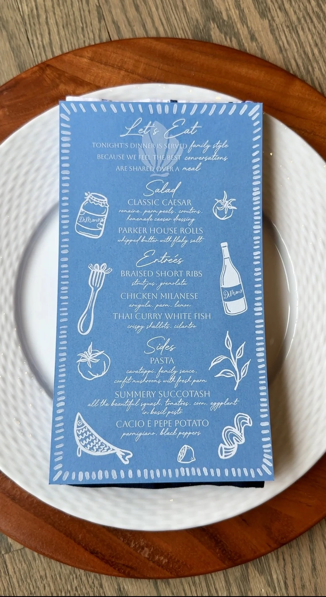 Blue dinner menu with handwritten-style text and white illustrations on a white plate, placed on a wooden table.