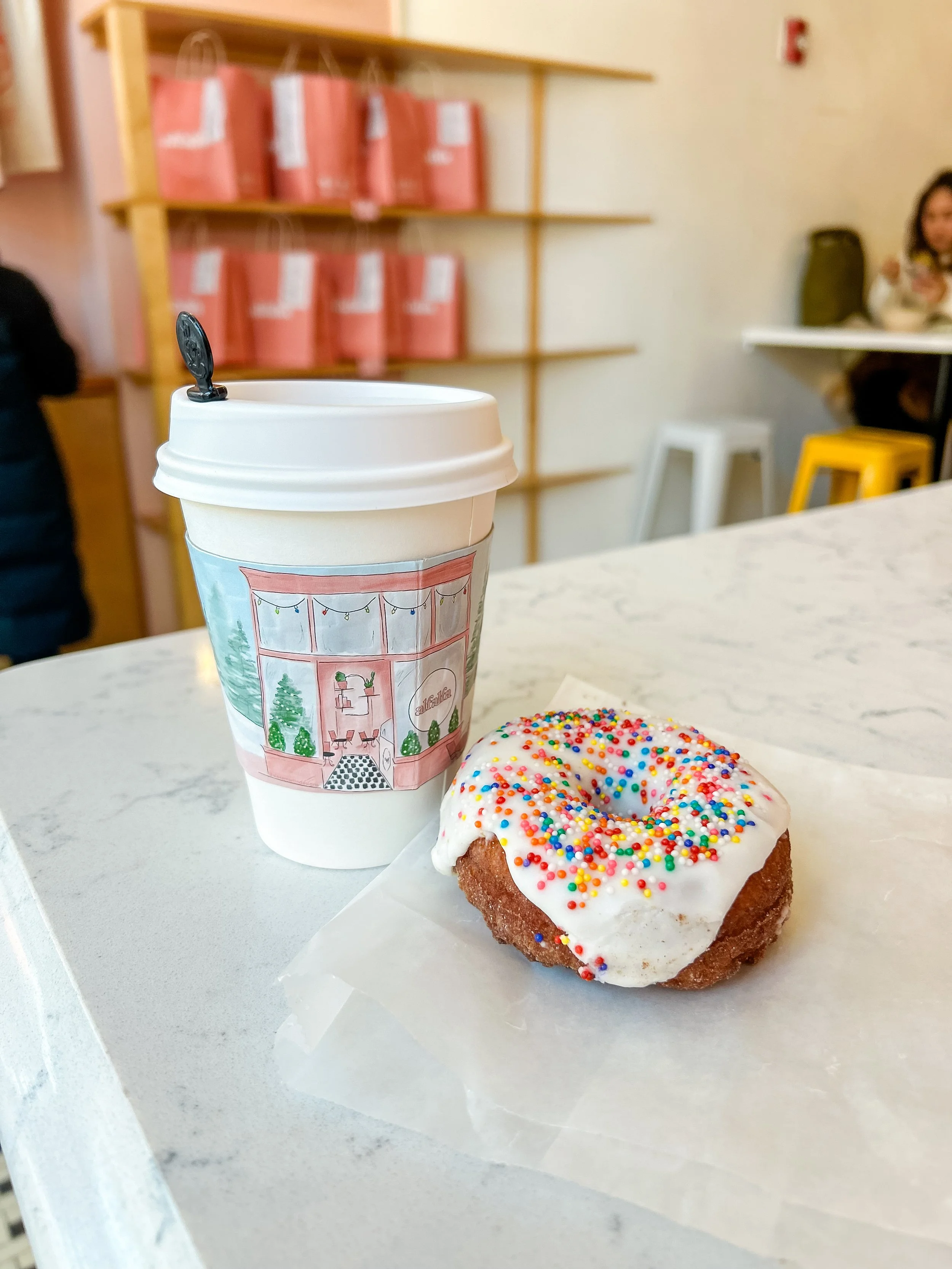 To-go coffee cup with a festive illustration of a window with string lights, a Christmas tree, and a cozy interior, alongside a iced donut with white icing and multicolored sprinkles on a white marble table in a cafe.