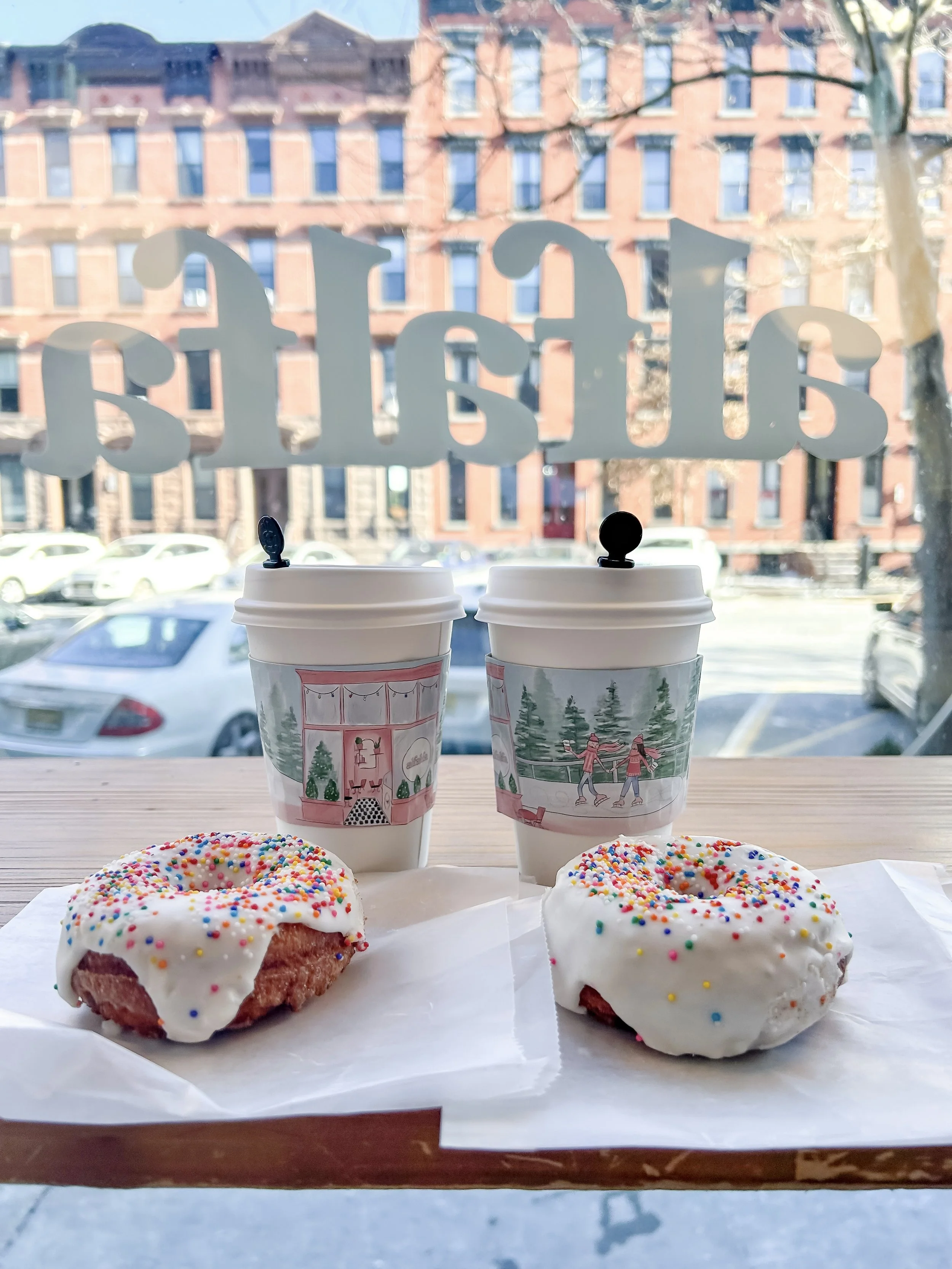 Two donuts with white icing and colorful sprinkles on parchment paper, two cups of coffee with holiday-themed sleeves, with a city view through a window with the word 'Bake' written on it.