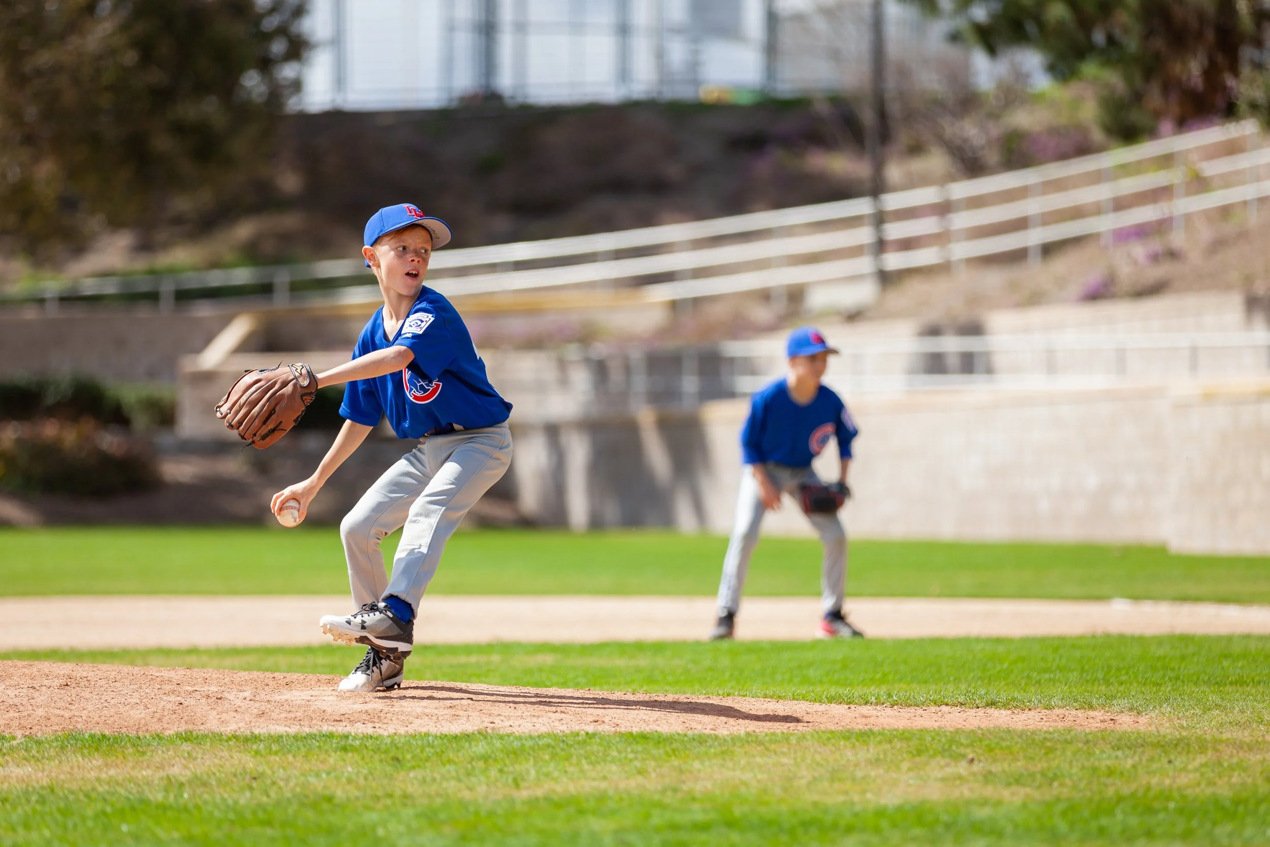 Little League Baseball pitcher in motion on pitching mound with first base player in the background.
