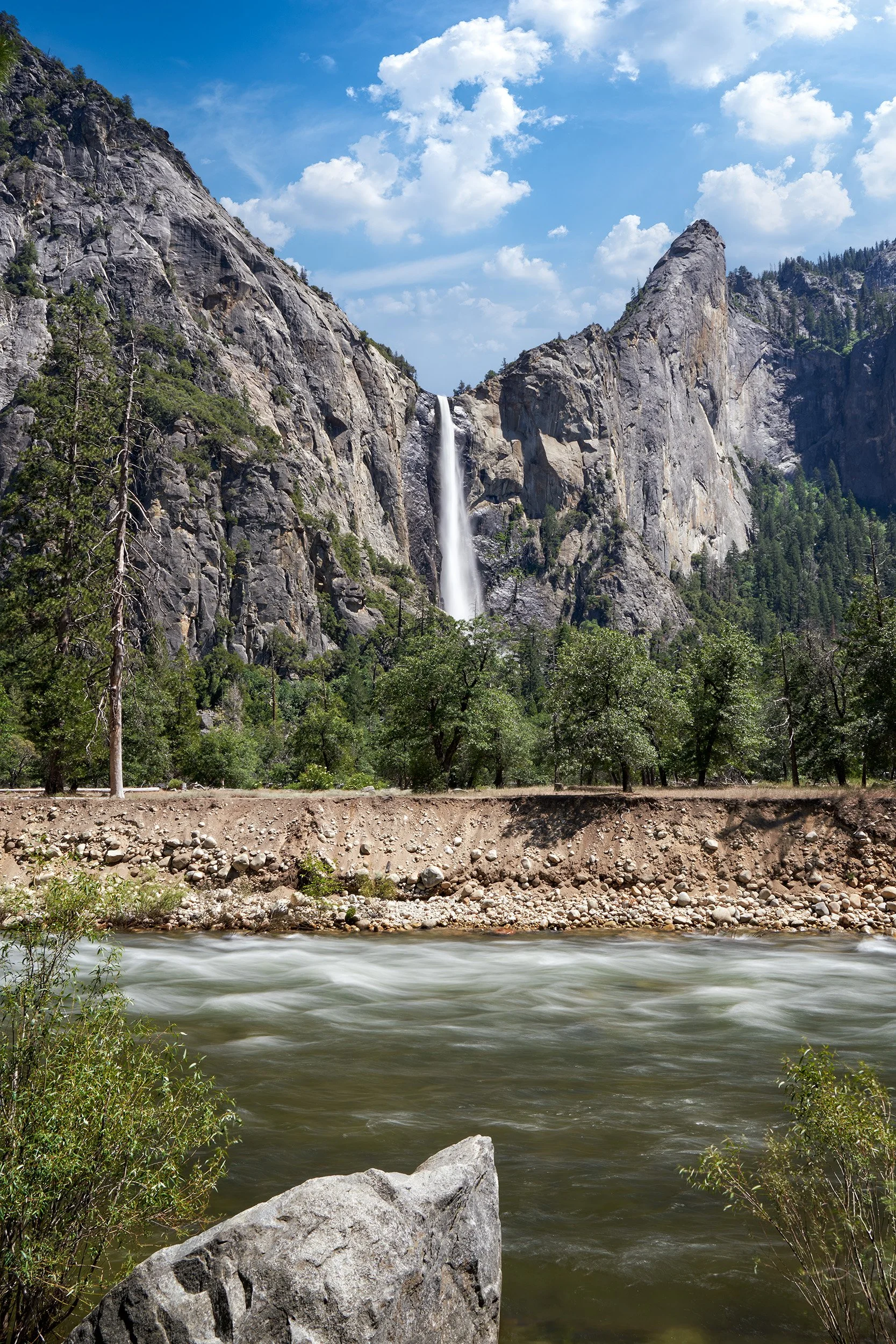 A waterfall cascading down a rocky mountain into a river with flowing water and green trees in the foreground, under a partly cloudy sky.