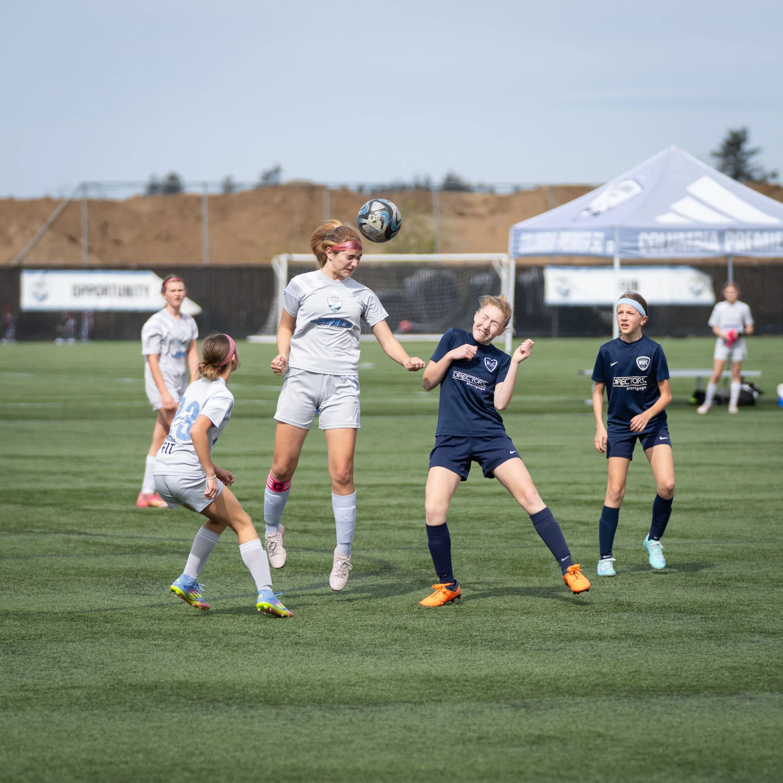 Young girls playing soccer on a grassy field during daytime, with a soccer ball in mid-air and some players watching, under a blue sky with a tent and fencing in the background.