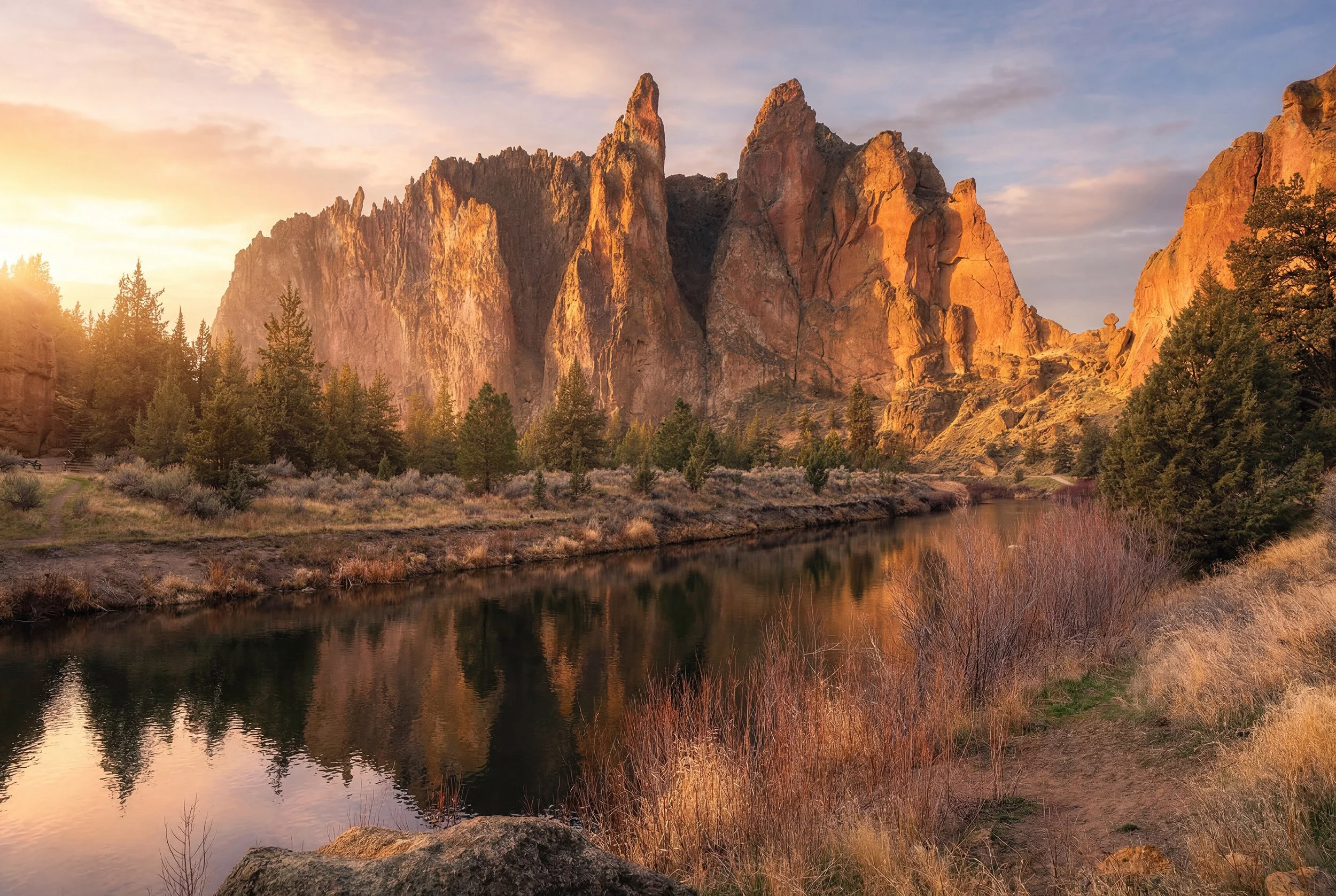 Morning light on Smith Rock State Park showing a rugged mountain with a river in the foreground and trees along the riverbank.